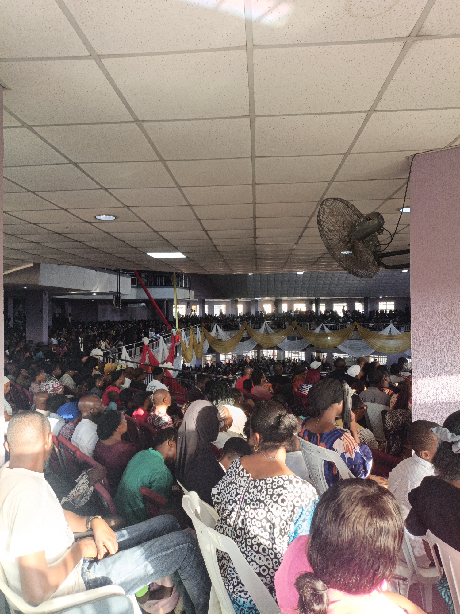 The image captures a large gathering of people, likely in a community hall or place of worship, within Sabon Gari West, Nigeria. The interior is illuminated by natural light streaming in from windows and by overhead lighting. The ceiling is a tiled drop ceiling with light fixtures. A large industrial fan is mounted on the wall near the ceiling, indicating the need for ventilation. The attendees, predominantly adults, are seated in rows of chairs, facing towards a common focal point that is not visible in the frame. Many individuals are looking upwards or towards the front, suggesting they are engaged in an event, possibly a service or a meeting. The attire of the people varies, with many wearing casual clothing. In the background, decorative drapes in gold and white are visible, suggesting a festive or formal occasion. The overall scene conveys a sense of communal gathering and participation.