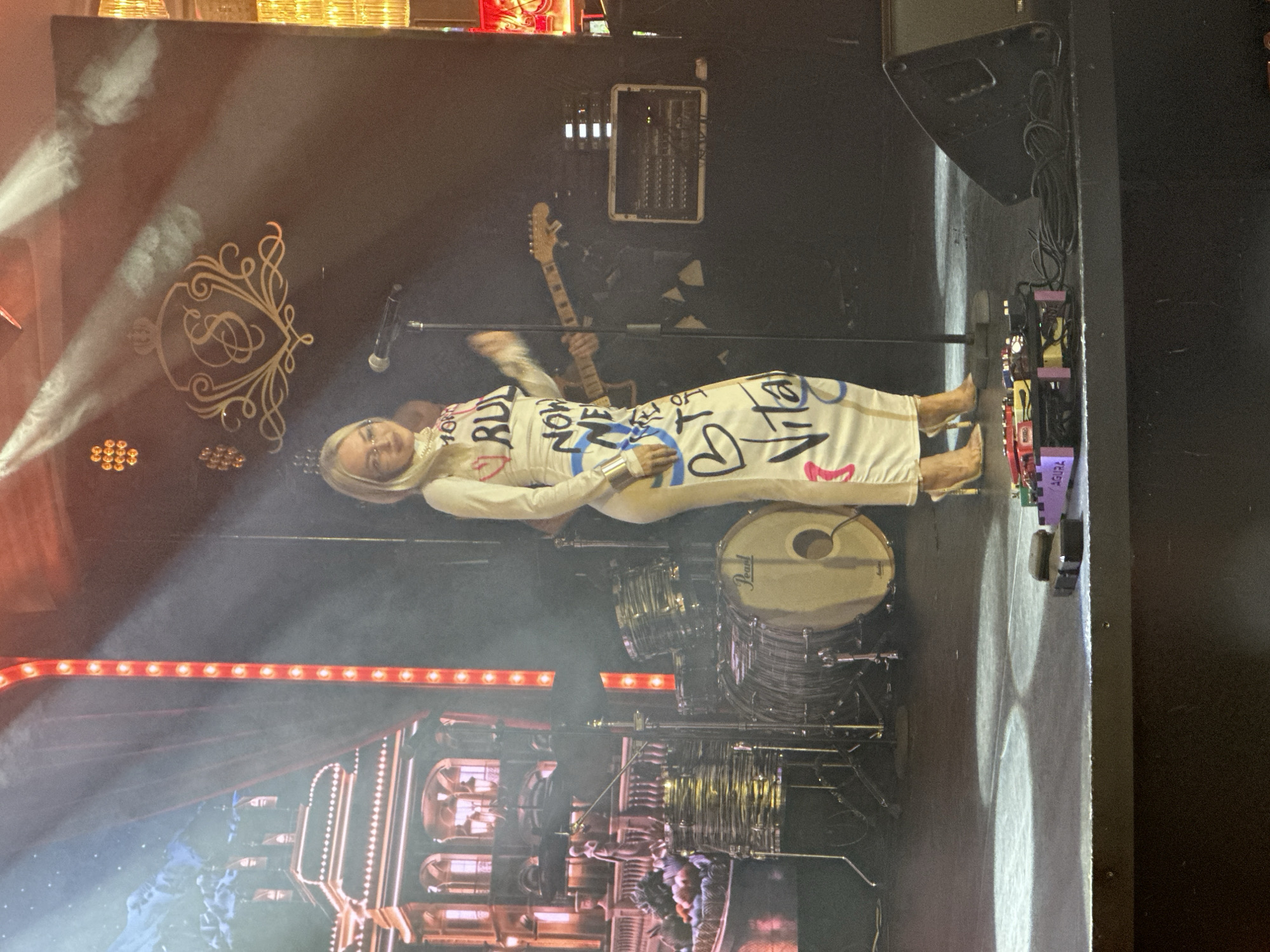 The image depicts a woman on a stage in Estosadok, Russia, likely at a music venue. She is wearing a long, white, form-fitting dress adorned with black and blue graffiti-style lettering. The woman has blonde hair and is wearing glasses and high heels. She is standing in front of a microphone and appears to be performing or about to perform. Behind her, a drum set, a guitar, and sound equipment are visible. The stage is lit with spotlights, and a backdrop featuring a depiction of a grand building and mountains is visible on the left. The overall scene suggests a live musical performance in an indoor setting.