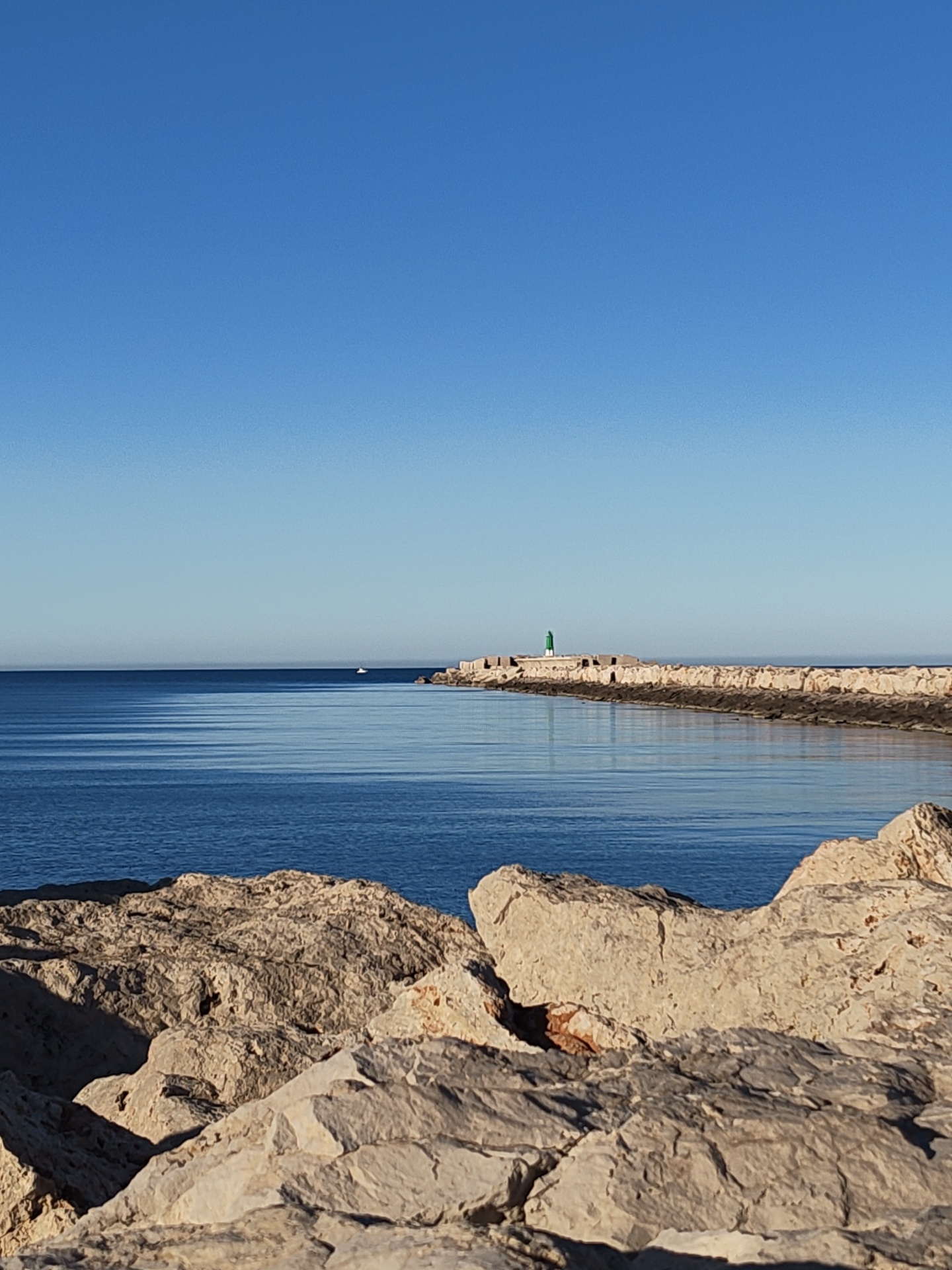 The image captures a tranquil coastal scene in Dénia, Spain. In the foreground, a rocky shoreline features light-colored, weathered stones, their surfaces marked by fissures and shadows. The calm, blue sea stretches horizontally across the middle ground, exhibiting gentle ripples and reflections. On the horizon, a long, man-made breakwater extends into the sea, culminating in a small, green-topped lighthouse. The sky above is a clear, gradient blue, transitioning from a deeper hue at the top to a lighter shade near the horizon. A small boat is visible far off in the horizon, between the breakwater and the shoreline. The overall atmosphere is one of peace and natural beauty.