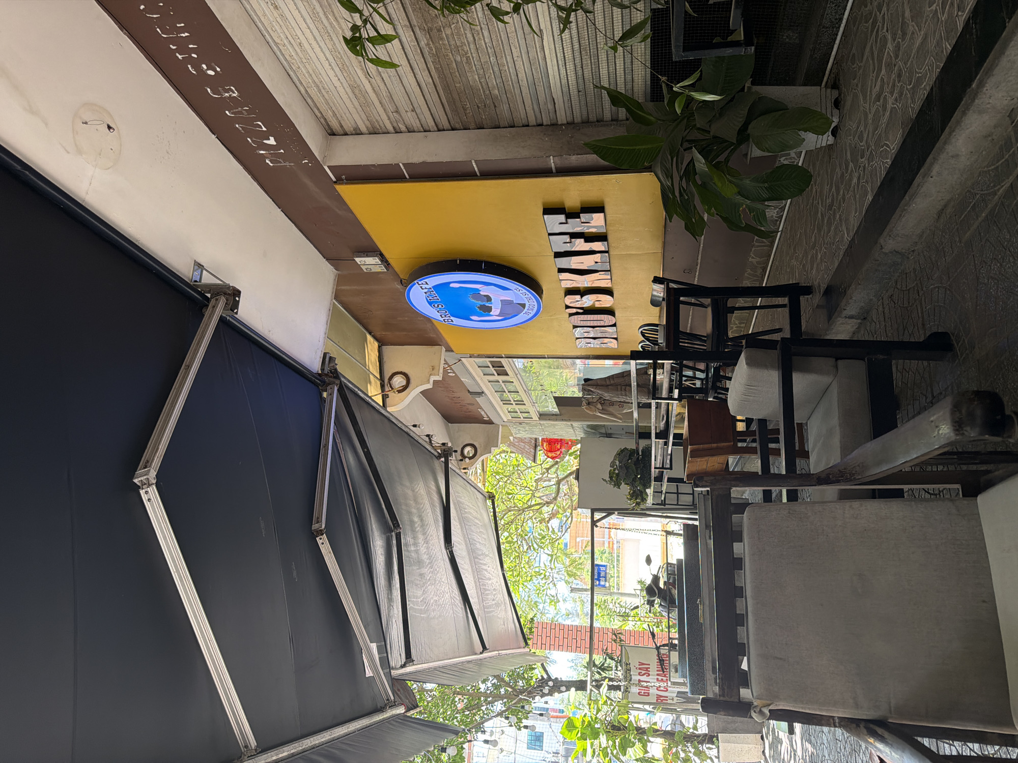 The image shows an outdoor seating area of a café or restaurant, likely in Đà Nẵng, Vietnam. A black awning partially covers the area, with visible supporting metal frames. Above the awning, faded text reads "PIZZAS STEL" along the wall. A yellow overhang is present with the café's name, "BROS KAFE," displayed in large block letters, and a circular sign with the café's logo.
Several chairs with light-colored cushions are visible, arranged as if for customers. A sign on a brick wall mentions "GIẶT SÂY RY CEA," indicating a possible laundry or cleaning service nearby. In the background, through the outdoor seating area, a street scene is visible with vegetation and buildings, indicating a typical urban environment in Vietnam.
