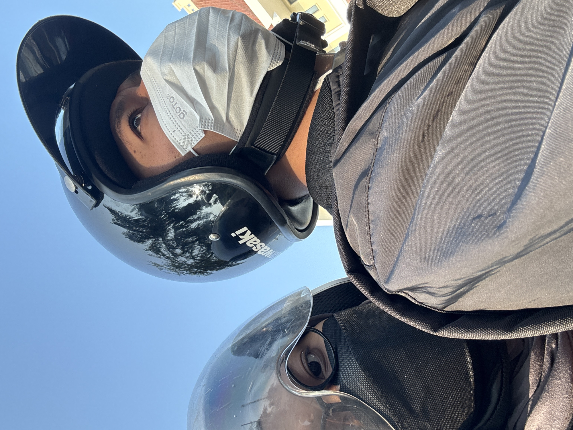 The image shows two people wearing helmets and face masks, likely on a motorcycle, under a clear blue sky. The person on the right, in the foreground, wears a black helmet with the "Kawasaki" logo visible, a white "GOTO" branded face mask, and a dark jacket. The person on the left, partially visible and slightly out of focus, wears a silver helmet with a clear visor and a black mask. The reflection on the silver helmet shows palm trees. In the background, a building with light-colored walls is visible. The lighting suggests it is daytime. The context implies they are in a city, potentially West Jakarta, Indonesia, given the presence of palm trees and the style of the building, common in tropical urban environments.