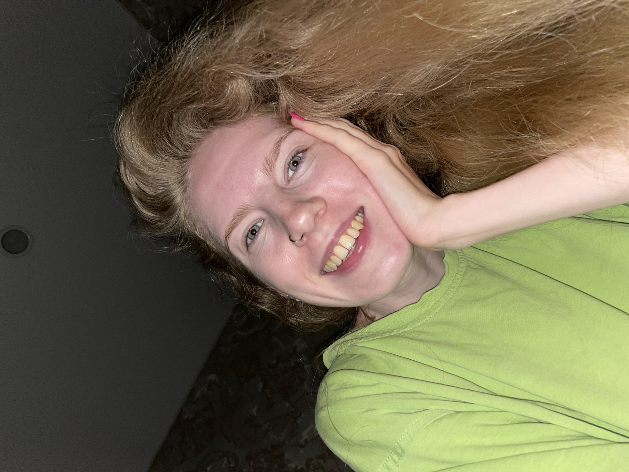 A young person, likely female, with long, voluminous, wavy light-brown or blonde hair, smiles broadly into the camera, tilting their head to the right. Their face is illuminated, highlighting light blue-grey eyes and a silver hoop nose ring on their right nostril. One hand, adorned with bright pink nail polish on the thumb, rests gently on their cheek and temple. They are wearing a vibrant lime green t-shirt, which has visible fabric folds and seams.
The individual's smile reveals their teeth, which have a yellowish tint. The background is predominantly dark, suggesting an indoor setting, possibly at night or in low light, with artificial illumination on the subject. To the lower left and behind the head, a dark patterned surface, resembling wallpaper or fabric, is visible. In the upper left corner, a dark, rectangular electronic device, possibly a tablet or phone, with a circular camera lens, is visible, suggesting the image is a selfie taken with this device. The overall mood conveyed is cheerful and relaxed. The setting is indoors, in Izhevsk, Russia. No visible text is present.