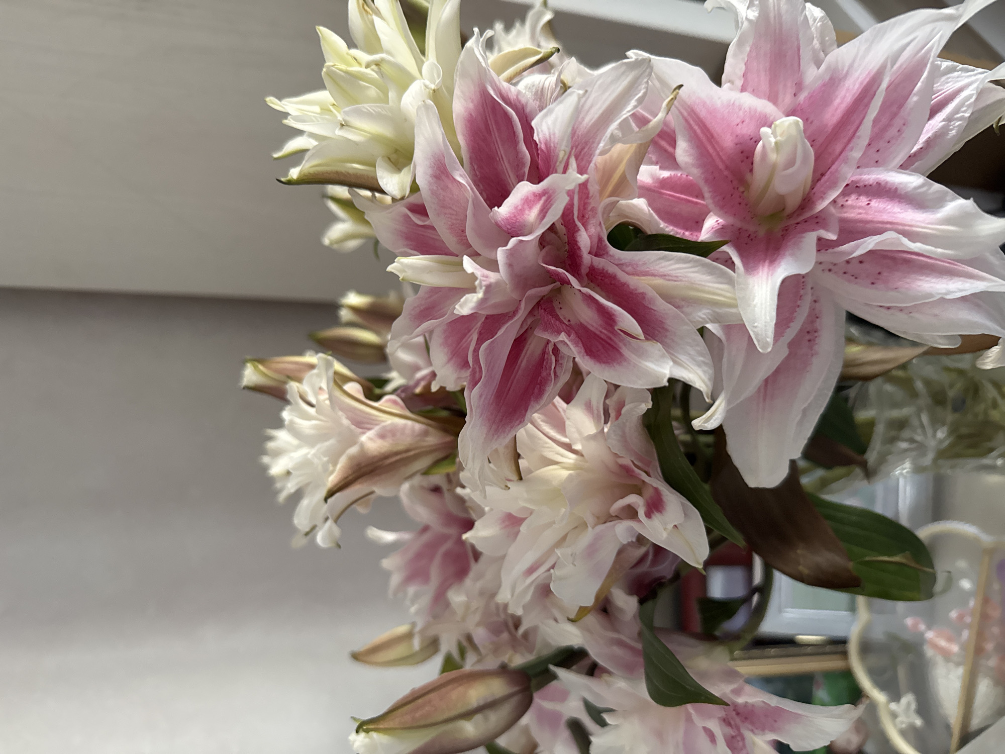 The image shows a close-up of a bouquet of double-petaled lilies, with a dominant color of pink and white. The lilies are in various stages of bloom, with some fully open and others still in bud. The background is a plain, light-colored wall, suggesting an indoor setting. There is a hint of a glass vase and some decorative items on a surface below the flowers, but these are out of focus and do not provide any specific details. No people are visible in the image. The image does not contain any information that would allow for the determination of its location within the city of Almaty, Kazakhstan.