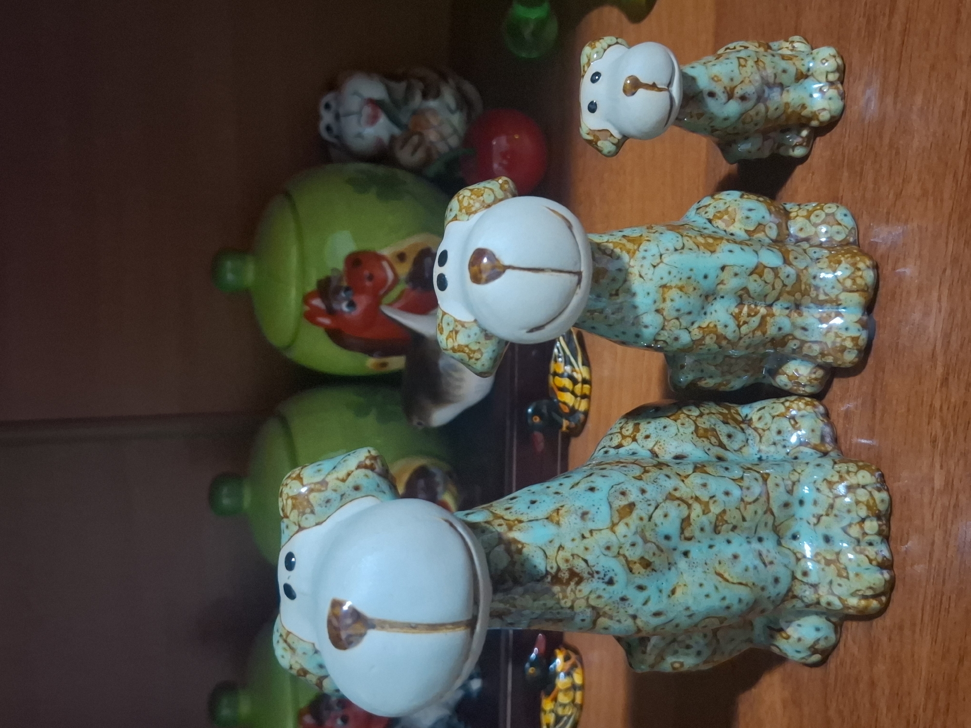 The image shows a collection of ceramic figurines on a wooden surface, likely a shelf or table. In the foreground, there are three dog figurines of varying sizes, all with light blue and brown speckled glaze. They have large heads with floppy ears, black dot eyes, and brown noses. Behind the dog figurines, there are two large green ceramic jars with lids. On the left jar, a red and brown horse figurine is visible. To the right of the jars, a white and brown rabbit figurine is depicted. Further to the right, a red sphere, possibly an apple figurine, is present. In the background, more ceramic objects are partially visible, including what appears to be a green bottle. Two small yellow and black duck figurines are placed on the wooden surface in front of the large dog figurine. The lighting suggests an indoor environment, possibly a home or shop. No specific location in Almaty, Kazakhstan can be determined from the image.
