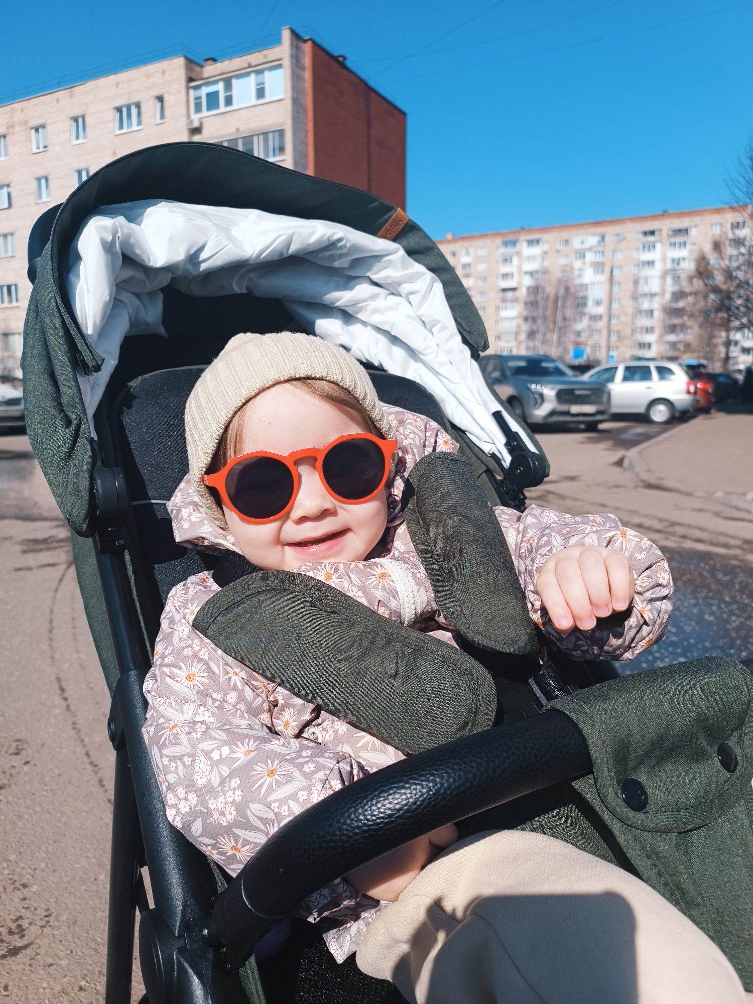 A toddler wearing a beige beanie and red sunglasses sits in a dark green stroller. The child wears a patterned jacket and is secured by dark green straps. Behind the stroller, a paved street is visible, with parked cars and apartment buildings in the background. The location is Izhevsk, Russia, as indicated by the architectural style of the buildings. The day appears sunny and clear.