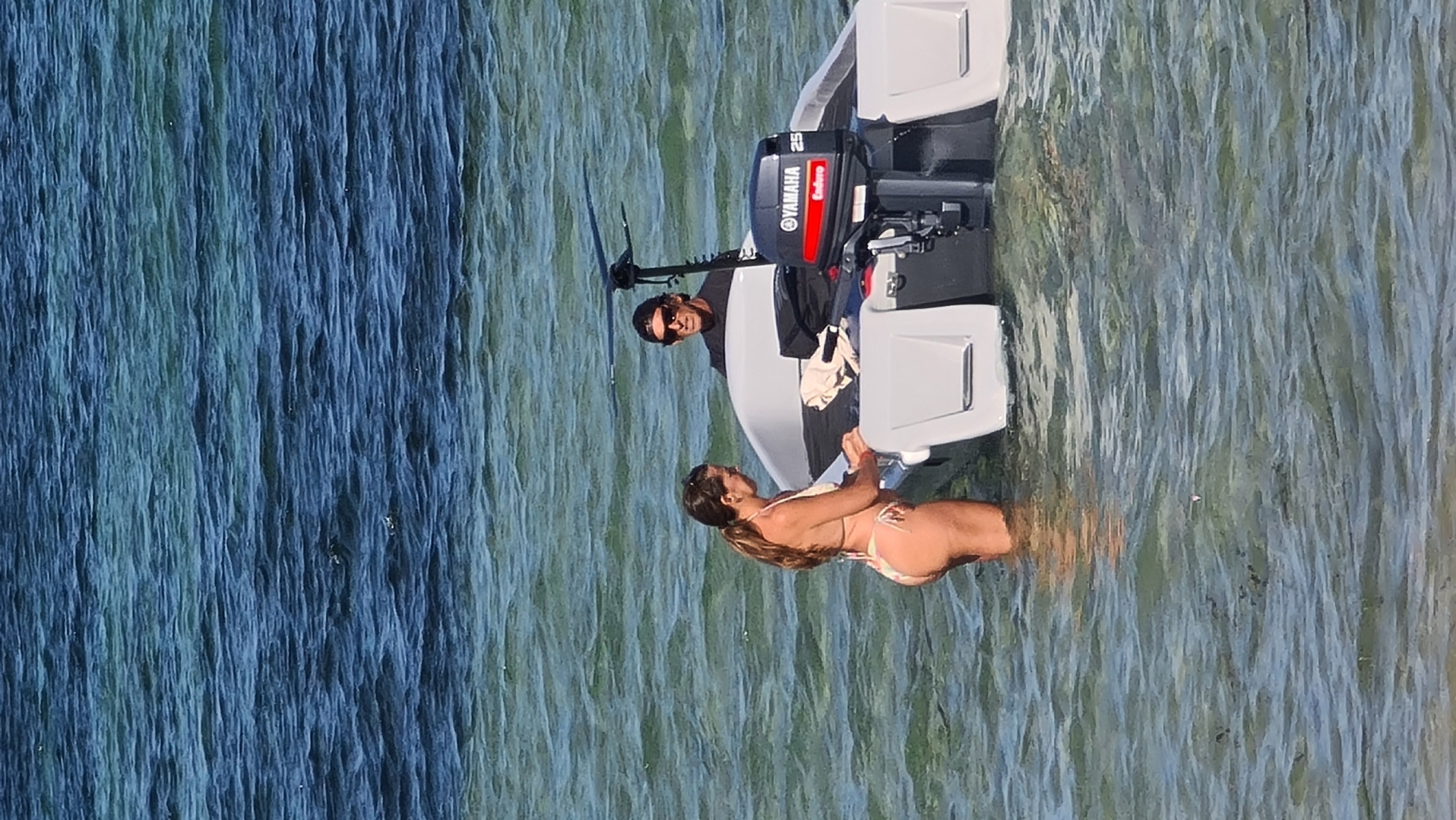 The image shows two people in shallow, clear ocean water near the shore. A woman in a patterned bikini is standing in the water, facing a small, white boat with a Yamaha outboard motor. She is reaching towards the boat. A man wearing sunglasses and a black t-shirt is seated in the boat, looking towards the woman. The water has ripples and the bottom is visible in some areas, suggesting shallow depth. The background consists of deeper blue ocean water. The scene appears to be a casual outing on the water, possibly in Denpasar, Indonesia, given the clear water and tropical appearance.