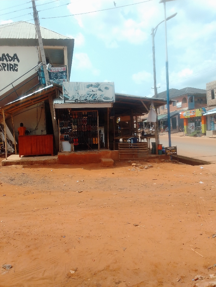The image shows a small, open-air shoe shop in Auchi, Nigeria, at the intersection of two dirt roads. The shop is located at coordinates 7.05 latitude, 6.263 longitude. The shop is made of wood and has a corrugated metal roof. It is brightly lit by the sun, and there are many pairs of shoes displayed on shelves. A single person stands at the counter, possibly a shopkeeper, with a red T-shirt on. The shop sign states "Windaabas' Empire to Less". The shop is facing a dirt road, where there is a metal streetlight pole in the background. On the other side of the road, there is a second building, a more modern, multi-story building with a blue door and a sign in white. The building appears to be closed. On the right side of the road, there is a smaller shop or stall, also facing the street. The sign on the stall says "Sally Beauty" in bold, white letters. The shop or stall has blue and yellow exterior. Both buildings appear to be closed and have a sign that reads "God Is Good". There are a few trees and other vegetation in the background. The sky is a light blue with a few white clouds. The dirt road in the foreground is a reddish brown color and appears dusty. The image is taken from a low angle, looking up at the shop.