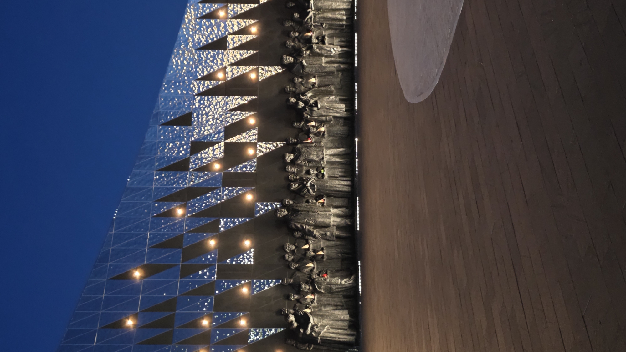 The image shows the Yad Vashem Holocaust Memorial Museum located in Jerusalem. The composition is captured from a low angle, emphasizing the museum's architectural design. A section of the building's exterior wall is visible on the left, featuring a geometric design composed of reflective triangular panels. There are lights positioned within the triangular cutouts. Adjacent to the wall is a dark, vertical display of bas-relief sculptures depicting a line of figures. To the right of the figures, the scene opens to a wooden floor. The sky is blue.