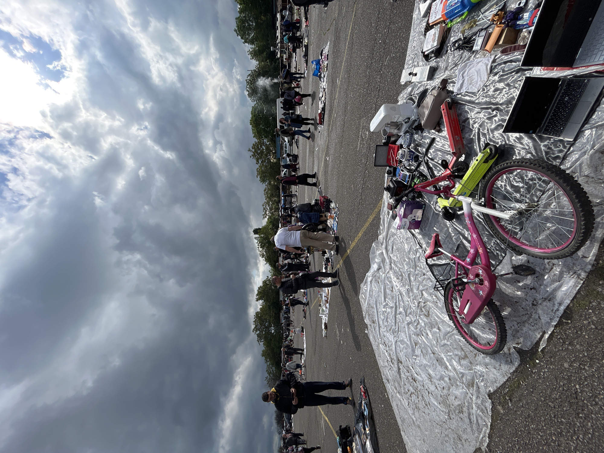 The image depicts a flea market likely in Sofia, Bulgaria, as evidenced by the architecture and Cyrillic script possibly present on some objects. The market is set in a paved area, possibly a parking lot, with numerous vendors displaying their goods on blankets and tables. A large crowd of people is present, browsing and interacting with the merchandise. The goods on display are varied, including electronics, bicycles, household items, and clothing. The sky is overcast with heavy clouds, suggesting the possibility of rain. A pink bicycle is prominently displayed on a tarp in the foreground.