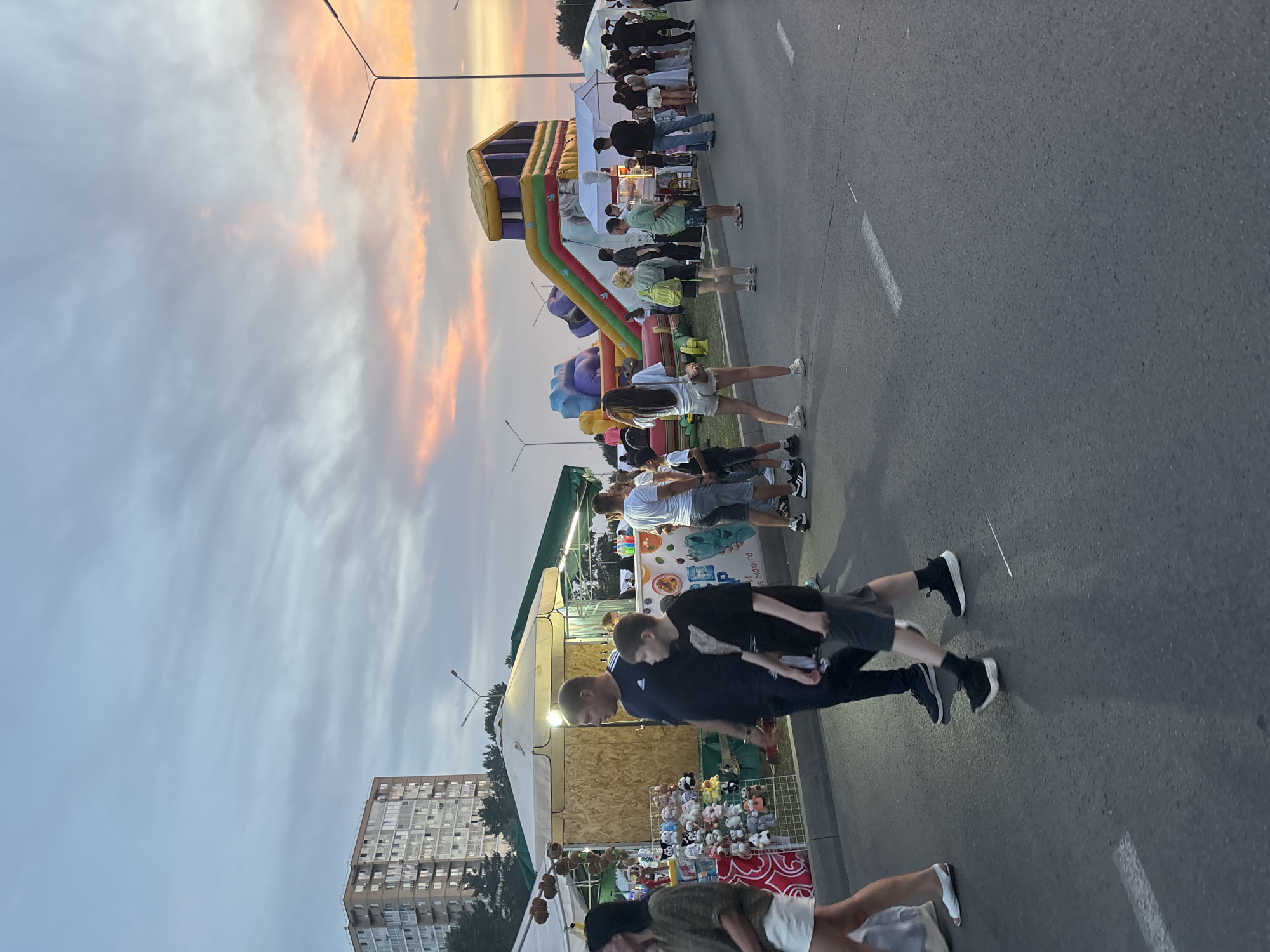 The image depicts an outdoor market or festival scene at dusk in Naberezhnye Chelny, Russia. A multi-story apartment building is visible in the background, along with a street lamp and a vibrant, colorful sky with pink and orange hues from the sunset. In the foreground, a paved road is lined with various stalls and attractions. People are walking along the road, suggesting a lively atmosphere. Among the stalls, there is an inflatable playground with a slide, several white tents or canopies, and displays of merchandise, including stuffed animals. The people in the image are dressed in casual clothing, indicative of warm weather. The overall impression is a community gathering or event taking place in an urban setting during the evening.