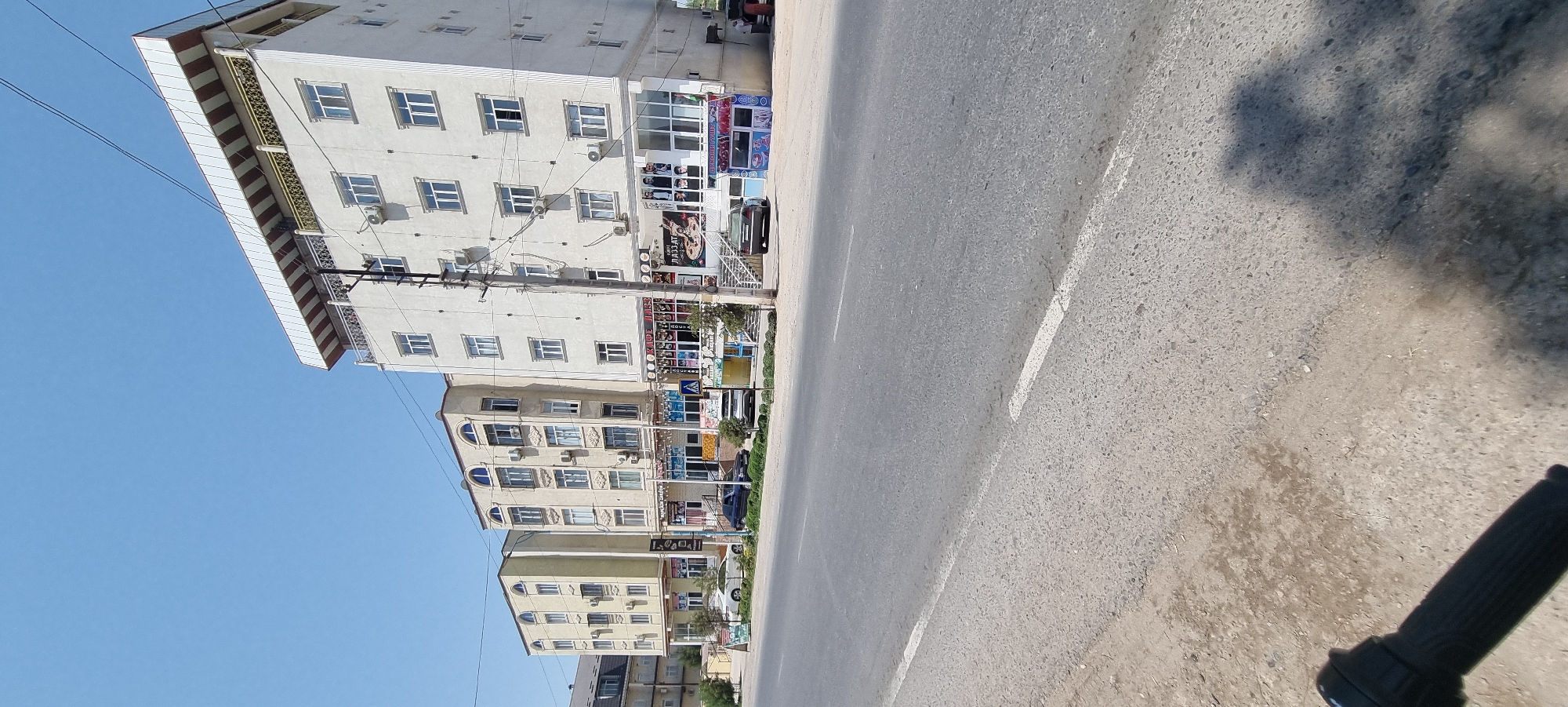 The image shows a street scene in the city of Ismoili Somoni, Tajikistan. On the right side of the street are several multi-story buildings with commercial establishments on the ground floor. The buildings have white and beige facades, with multiple windows and air conditioning units. The commercial spaces appear to be shops or restaurants, with signage visible in Cyrillic script. One sign reads "KAФE ЛAЗЗАТ". There are people visible on the sidewalk in front of one of the buildings, and a few cars are parked on the street. The street itself is paved and appears to be a main thoroughfare. The sky is clear and blue, suggesting a sunny day. A power pole with electrical wires runs along the street in front of the buildings. In the foreground, a portion of a black bicycle handlebar is visible, indicating the photo was taken from a bicycle.