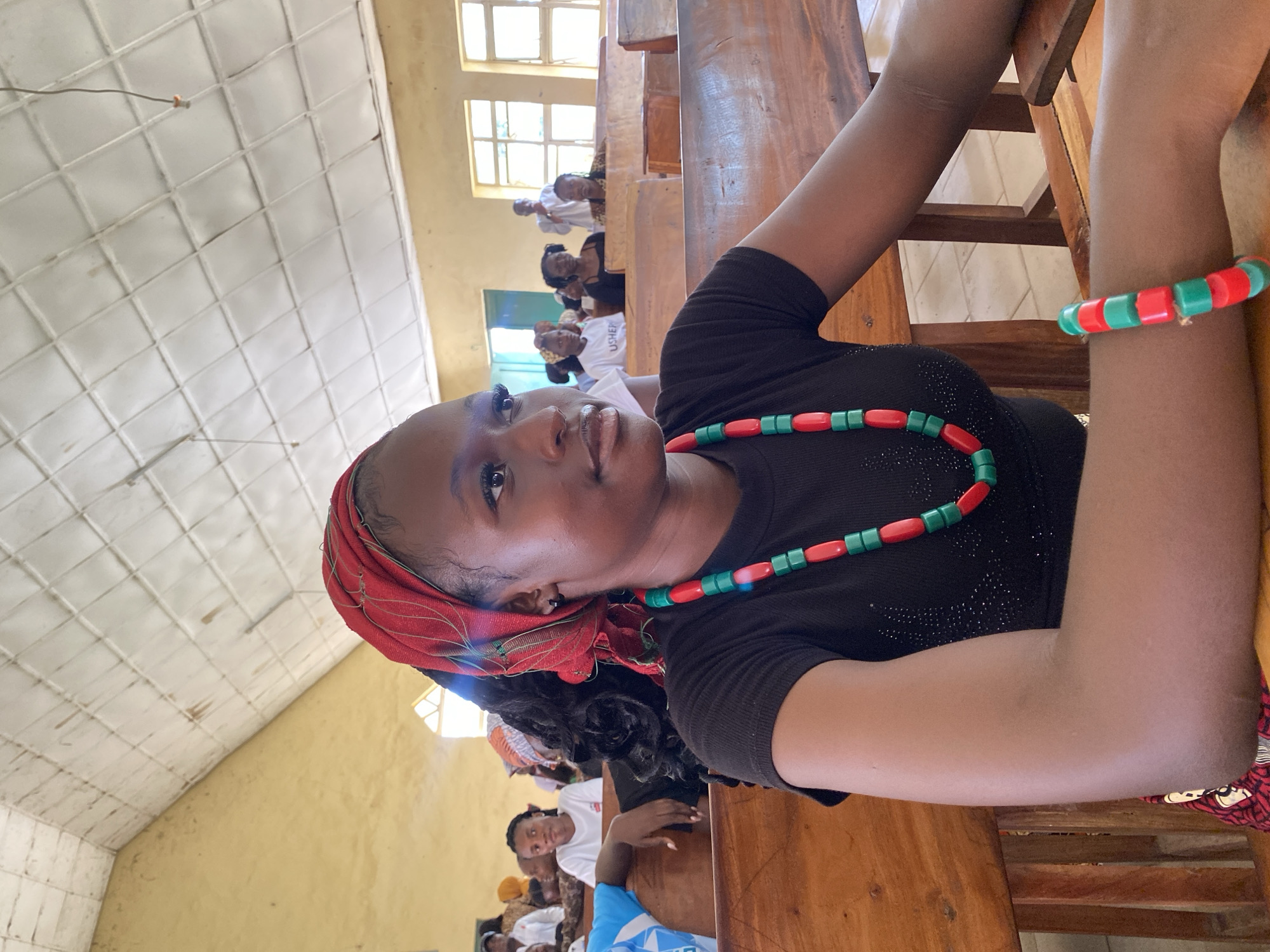 The media file depicts a gathering of people in what appears to be a community hall or church in Dutsen Kato, Nigeria. The image is oriented vertically, with the primary subject, a young woman, prominently featured in the foreground. She is wearing a black top adorned with subtle sparkle details, a red headscarf, and a beaded necklace and bracelet in alternating red and green colors. Her gaze is directed slightly upwards and to the left, with a neutral expression.
Behind her, a row of other individuals are seated at wooden benches. Some are looking towards the camera, while others appear to be engaged in conversation or observing something. The setting suggests an indoor event, possibly a service or a community meeting, given the presence of multiple people seated in rows. The room has a high, white, textured ceiling and yellow walls. Windows with visible panes are present, suggesting natural light is entering the space, though the time of day is not explicitly discernible.
One individual in the background, wearing a white t-shirt, has text on it, but it is partially obscured and unreadable. Another person in a white garment is visible further back. The overall atmosphere seems calm and communal. The wooden furniture and ceiling design are consistent with local architecture or community building styles.