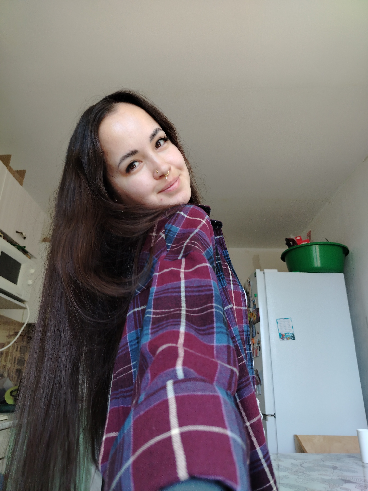 A young woman is in what appears to be a kitchen. She has long, dark hair and is wearing a plaid shirt of burgundy, blue, and white. She is looking at the camera with a slight smile. In the background, there's a white refrigerator with magnets on it, a green bowl on top of the refrigerator, and white kitchen cabinets. The location is the city of Noviy, Russia.