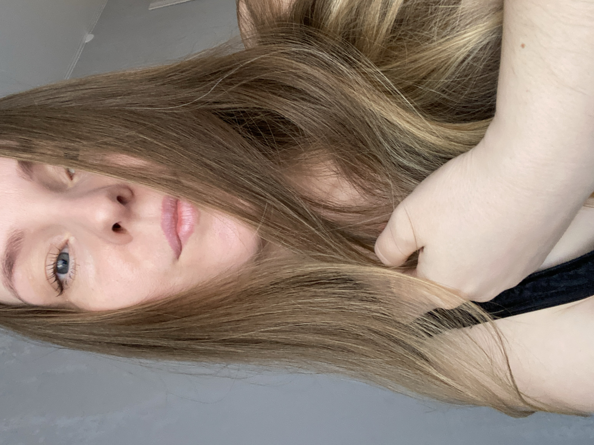 A close-up, angled view of a light-skinned woman with long, light brown hair. Her right hand is raised, possibly touching or holding her hair. She has light-colored eyes. A black article of clothing is visible near her shoulder. The background is a plain gray. There is no indication of the city of Noviy, Russia, or any other specific location.