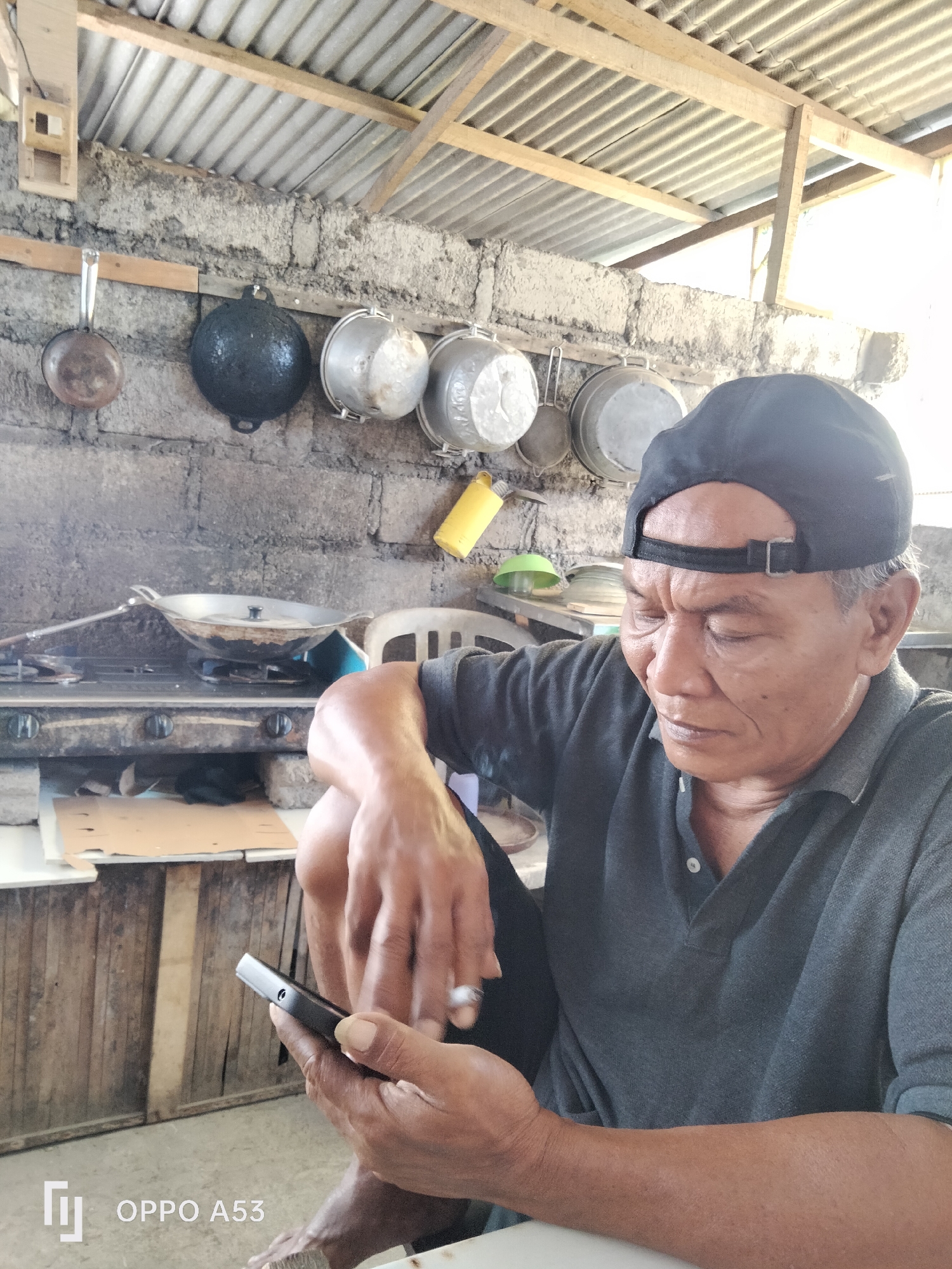 An older man with graying hair and a dark baseball cap is looking down at his mobile phone, which he is holding in his hands. He is wearing a dark gray polo shirt. The man appears to be in an outdoor kitchen area, with a corrugated metal roof supported by wooden beams overhead. The wall behind him is constructed from concrete blocks. Various cooking pots and utensils are hanging on the wall. A gas stove with a wok on it is visible in the background. The overall environment suggests a humble, possibly rural or semi-rural setting in Denpasar, Indonesia. The man's hands are visible, and he is holding a lit cigarette between his fingers.