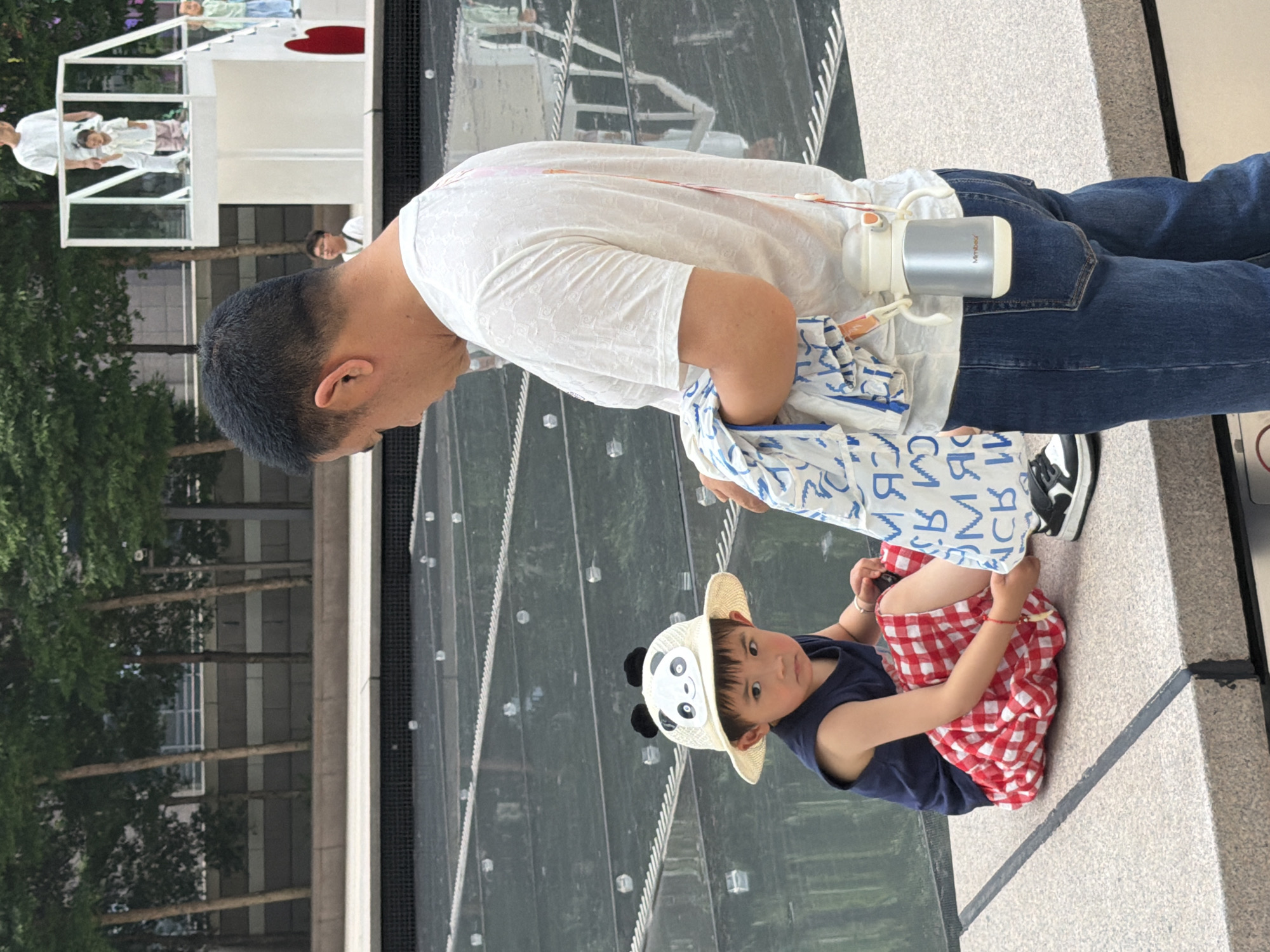 In the Yuzhong District of China, a man in a white t-shirt and blue jeans stands facing a young child. The man appears to have a pink-ish stain running down his back from his t-shirt. He is holding a white cloth with blue lettering. A metallic water bottle is clipped to his belt. The child, wearing a navy blue sleeveless shirt and a red and white checkered skirt, is seated on a stone ledge and is wearing a straw hat with a panda design. The child is looking towards the man. In the background, there are trees and a multi-story structure with people visible on it, suggesting an urban setting. The ground in front of the ledge appears to be a reflective surface, possibly water or glass.