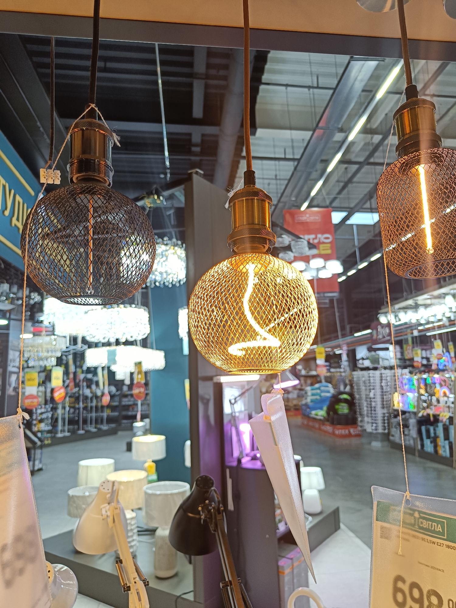 This media file shows a close-up view of three pendant light fixtures in what appears to be a retail store. The foreground features two spherical light bulbs encased in decorative metal cages, one with a dark, cage-like exterior and the other with a golden, intricate mesh. Both bulbs emit a warm, yellowish glow from their filament. A third light fixture, partially visible on the right, also has a similar decorative cage and illuminated bulb.
In the background, through a reflective surface that might be a window or a display case, a retail store interior is visible. Rows of other lighting fixtures, including chandeliers and table lamps, are displayed on shelves and racks. Some of these also appear to be illuminated. Signs with text are visible in the background, suggesting a sale or product information. One sign in the lower right corner has the text "СВІТЛА" (Svitla, meaning "light" in Ukrainian) and what appears to be a price, with the number "698" prominently displayed. The setting seems to be an indoor lighting store, likely during daylight hours, given the ambient light and reflections. There are no people visible in the image. The overall impression is a display of decorative and functional lighting products.