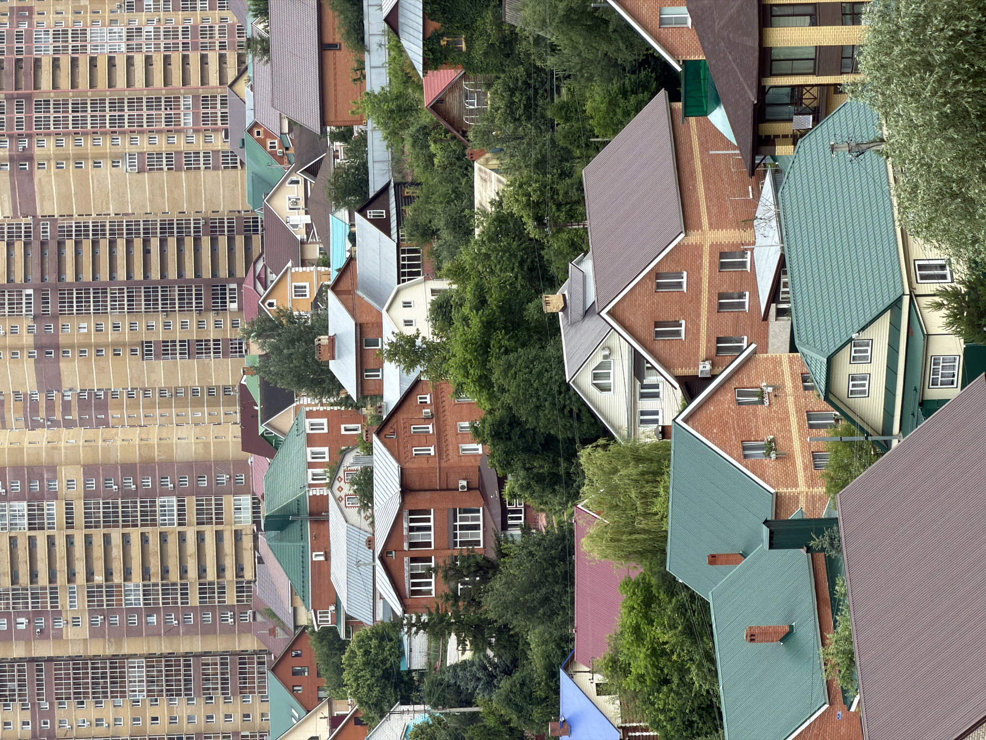 This is a high-angle, eye-level shot of a residential area, featuring a mix of modern apartment buildings and smaller, older houses. The apartment buildings on the left are tall and have a repeating pattern of windows and balconies, suggesting a dense urban or suburban environment. The houses on the right are varied in style, with brick and painted facades, pitched roofs in various colors like green, brown, and red, and are interspersed with lush green trees. Some of the houses have visible chimneys and air conditioning units. The overall impression is a densely populated area with a contrast between modern high-rise living and more traditional, individual homes. The lighting suggests it is daytime, and the presence of foliage indicates a temperate climate, possibly spring or summer. No people are visible in the frame, focusing the attention solely on the architecture and the natural elements. The image is vertically oriented and appears to be taken from a high vantage point, possibly a drone or a taller building, looking down and across the neighborhood. There is no discernible text in the image.
