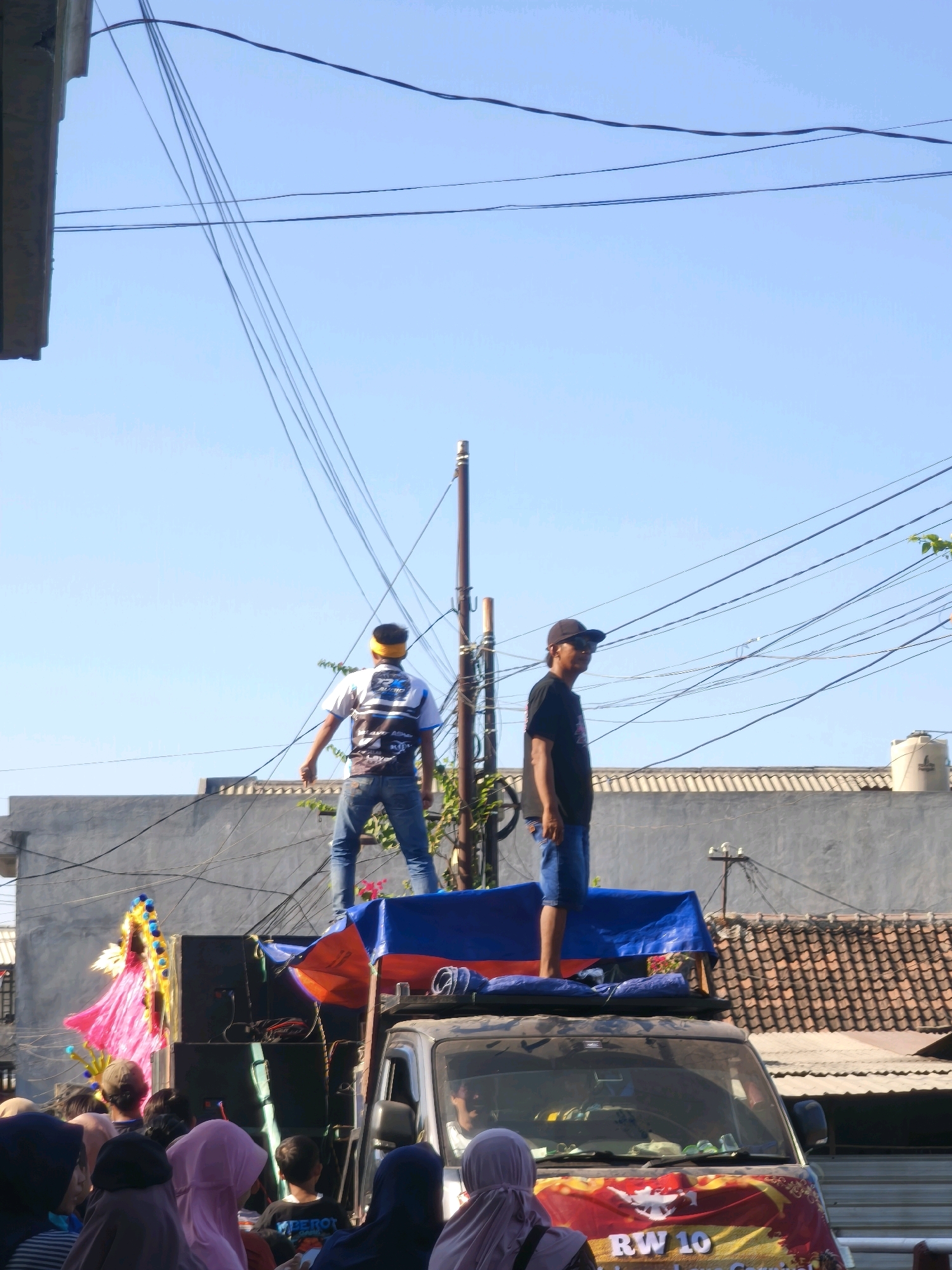 In the city of Kepuh Kiriman, Indonesia, on a bright, clear day, a street event is underway. Two men stand atop a vehicle, the one on the left wearing a white and blue patterned shirt with a yellow headband, and the one on the right in a black t-shirt and a black baseball cap. Below them, a crowd of people, many wearing headscarves, are gathered, looking towards a colorful float with a figure in a pink costume. The vehicle appears to be a speaker truck, with large black speakers mounted on its roof, draped with blue and orange tarps. A banner on the front of the truck reads "RW 10". The background features buildings with tiled roofs, a concrete wall, and a utility pole with numerous electrical wires crisscrossing the sky, indicating an urban or suburban setting. The overall scene suggests a parade or festival procession.