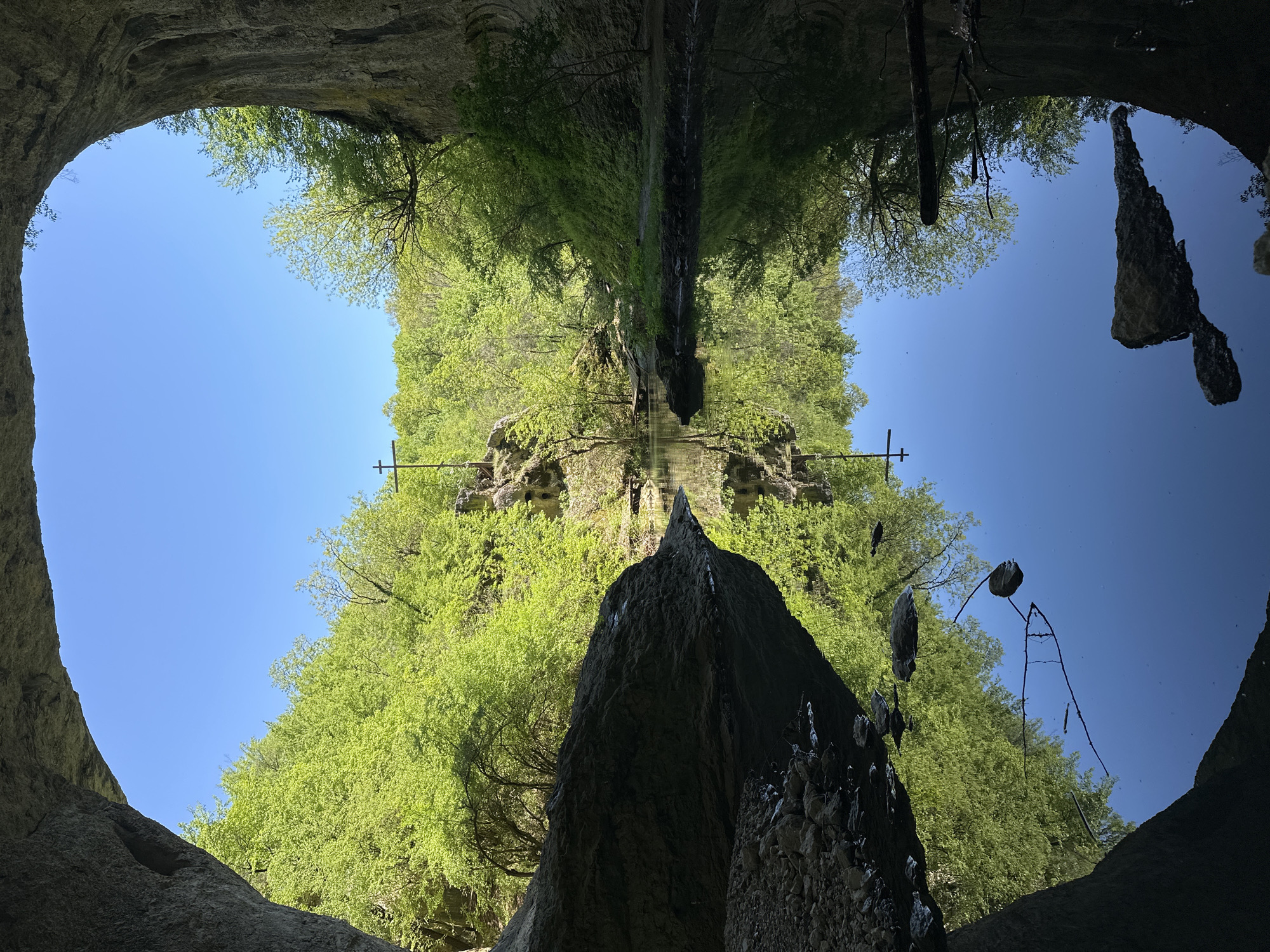 The image shows the Devetashka cave near the city of Lovech in Bulgaria, reflected in a pool of water. The cave entrance forms a rounded opening in the top portion of the image, revealing a clear blue sky beyond. Lush green vegetation covers the rock formations around the cave entrance. The reflection in the water creates a symmetrical effect, with the mirrored image appearing below the actual cave entrance. A large, dark rock formation is visible in the bottom foreground. A waterfall flows down the middle of the reflection. Two metal crosses or supports are visible to the left and right of the reflection. The water surface is mostly still, allowing for a clear reflection.