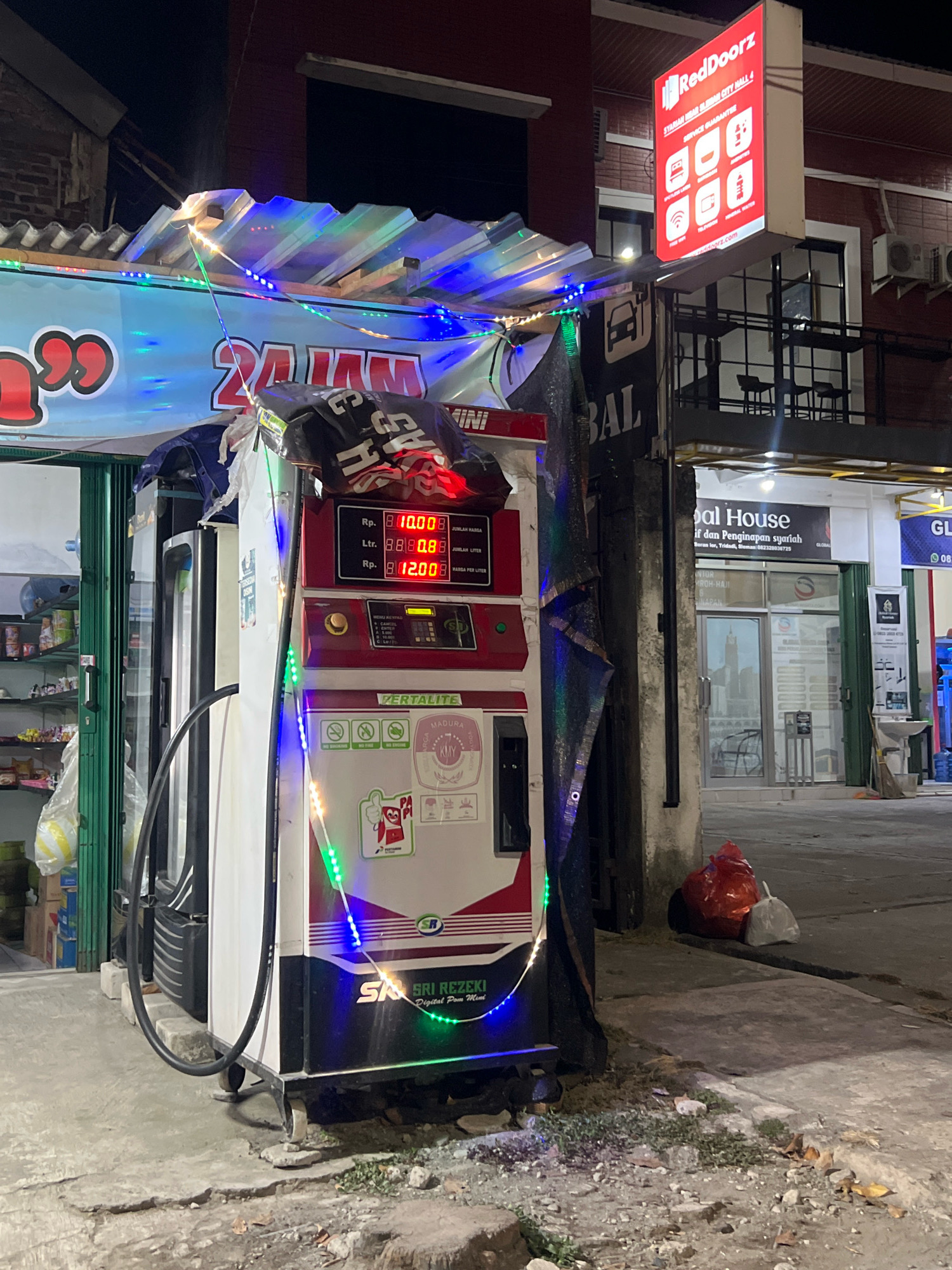 The image depicts a gas station and a RedDoorz hotel in the city of Sleman, Indonesia. In the foreground, a Pertalite gas pump is illuminated by colorful string lights. The pump displays fuel prices and quantities. To the left of the gas pump, shelves stocked with various items are visible behind a green metal gate. Behind the gas pump, a portion of a building with a corrugated roof is visible, also decorated with string lights. A RedDoorz sign is prominently displayed on the building to the right, advertising the hotel with icons representing services. The hotel's entrance is visible, with glass doors and windows. The environment is an outdoor commercial area, with pavement and debris on the ground. The time of day appears to be night, as evidenced by the artificial lighting and the darkness of the sky. No people are clearly visible in the image.