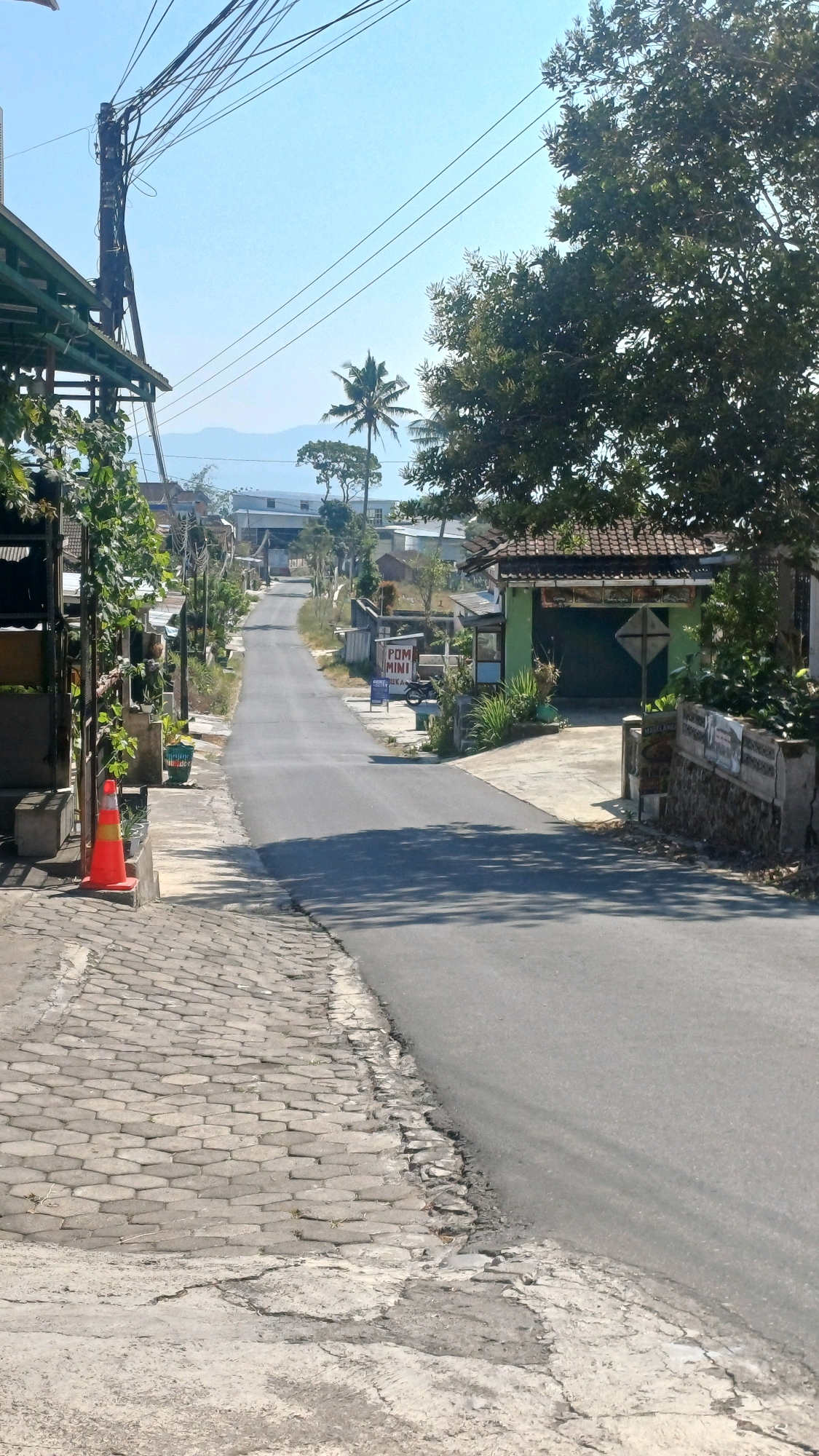 The image depicts a paved road winding uphill through a rural or semi-rural area, likely in Mungseng, Indonesia. The road is flanked by a cobblestone sidewalk on the left and a mix of concrete and asphalt on the right. Several buildings, some with tiled roofs and one with a green facade and a large garage door, are visible along the sides of the road. Vegetation, including trees and bushes, is present throughout the scene. A tall utility pole with numerous wires dominates the upper left portion of the image. In the distance, mountains and the sky are visible. An orange traffic cone is placed on the cobblestone sidewalk. A sign reading "POM MINI" is visible on the right side of the road. The overall impression is a bright, sunny day in a somewhat weathered, developed area.