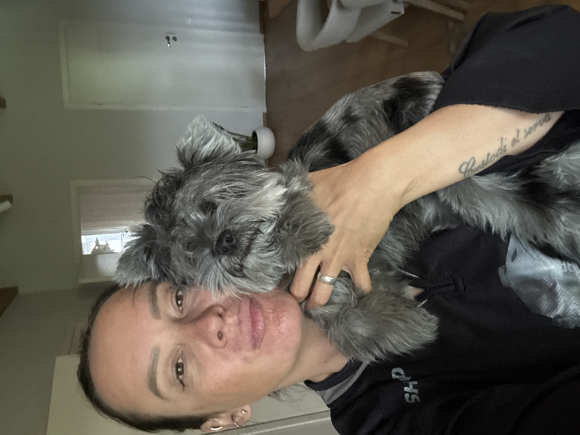 A selfie captures a woman holding a grey Schnauzer dog in her arms. The woman has light skin, brown eyes, and dark hair pulled back from her face. She is wearing a black shirt with white lettering partially visible. Her left arm is around the dog, and a tattoo is visible on her forearm. A silver ring is on her finger. The dog has a grey coat and a wiry beard. They are inside a room with white walls, a door, and a white chair. A window with light coming through is visible. The location is Chaikovsky, Russia.