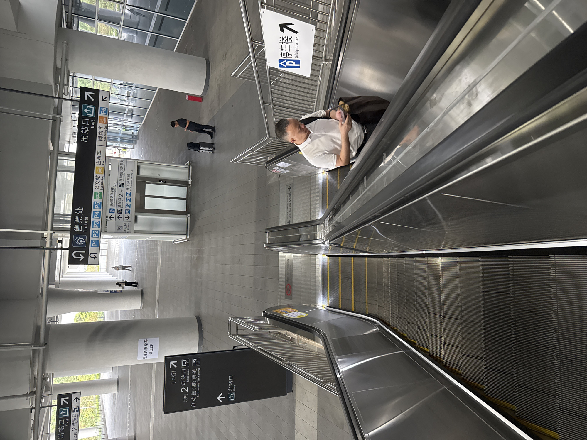 The image captures an escalator scene in a modern transportation hub, likely a train station or bus terminal, within Huizhou, China. In the foreground, a man wearing a white polo shirt and dark pants is descending an escalator, engrossed in his mobile phone. He is carrying a dark bag over his shoulder. The escalator itself is constructed of metal, with yellow safety lines along the edges.
Above and in the background, various signs indicate directions and services. One prominent black sign with white lettering points to "Tickets" (售票处) with associated platform numbers P1, P2, P3, P4, P5. Other signs direct towards "Bus Station" (公交站), "Taxi" (出租车), "Ride-hailing Car" (网约车), and "Parking Structure" (停车场). Another sign indicates "Entrance" (进站口) on the 2F level and "Automatic Ticketing" (自动售(取)票处), along with an "Exit" (出站口) sign.
The environment appears to be an indoor public space with large pillars supporting an elevated structure. Natural light filters in from large glass windows on the right. Two other individuals are visible in the mid-ground, one standing near a pillar and another walking away from the camera. Luggage, including a suitcase and a backpack, is present near a man standing in the background to the right of the image. The overall impression is one of a functional and well-signed transit facility.