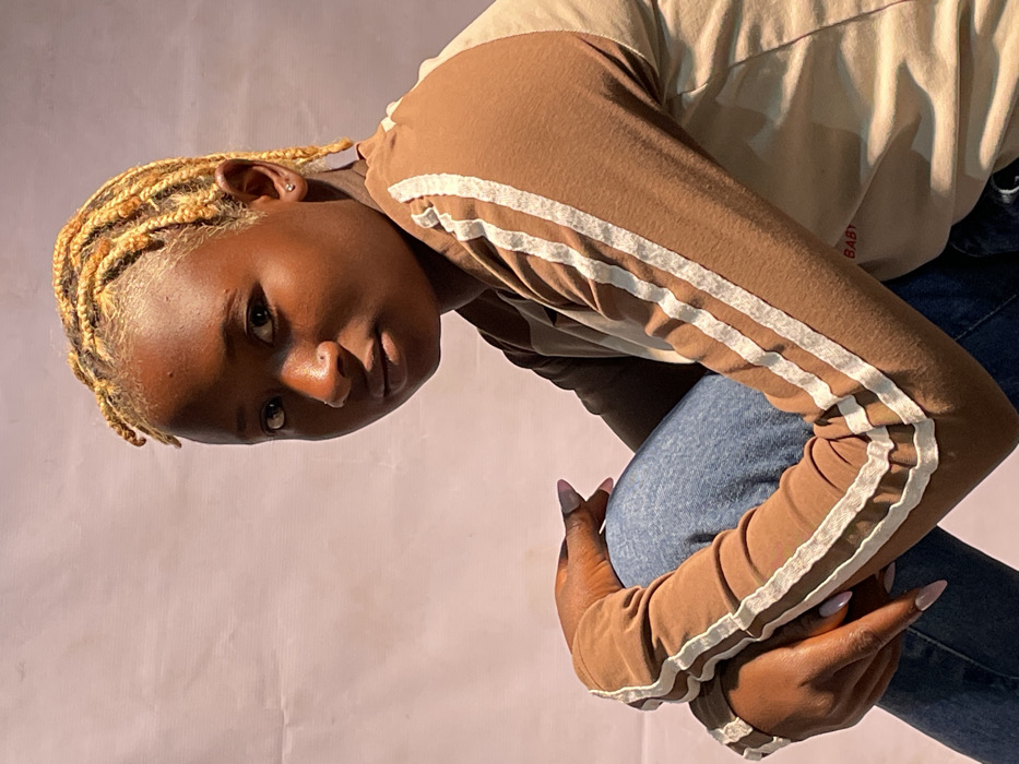 A young Black female, appearing to be in her late teens or early twenties, is depicted in a medium close-up, seated portrait against a plain, light pink/beige background exhibiting minor creases. She has dark skin and short, bleached blonde dreadlocks or braids. Her dark eyes are directed forward, towards the camera. A small silver stud is visible in her left nostril, and a similar stud earring is present in her left ear. She wears a two-toned long-sleeve shirt; the sleeves are brown with two vertical white stripes, while the torso is a lighter beige or light brown, featuring partial red text reading "BABY." She is wearing blue denim jeans. Her arms are bent, with her hands, which have long nails painted with light pink polish, resting on her knees. The setting is in the city of Jos, Nigeria. Soft, natural light illuminates the subject, casting subtle shadows.