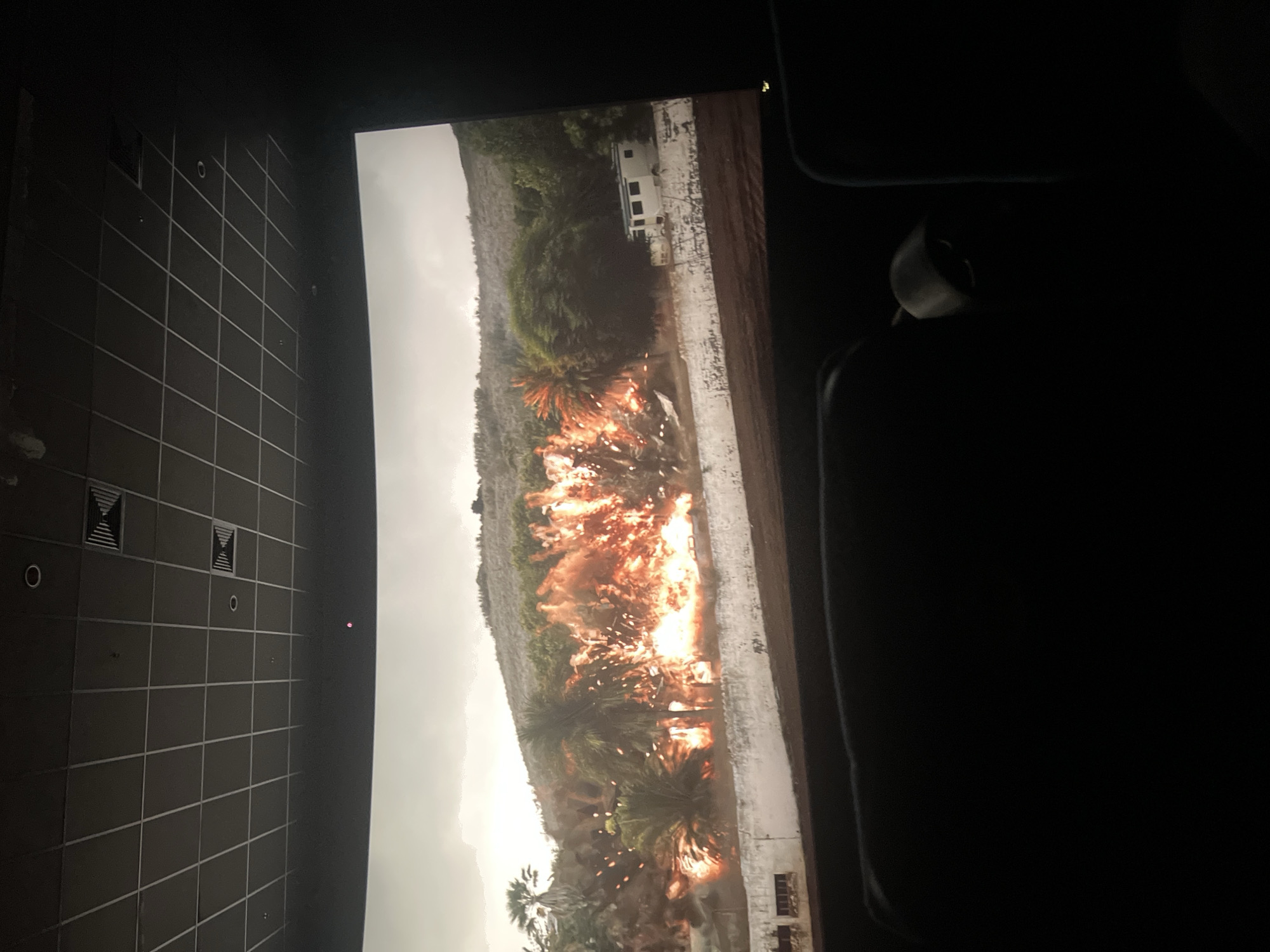 The image depicts a movie screen in a dark theater. The screen shows a scene of an explosion occurring in a natural landscape with palm trees and some buildings present. The explosion is the central focus, with bright orange and yellow flames. The surrounding environment appears to be somewhat overcast. Part of the theater's wall and ceiling is visible on the left side of the image, showing a grid-like pattern. A part of a theater seat is visible on the right side of the image. It is impossible to tell if the scene is in Krasnoyarsk, Russia, with only this image.