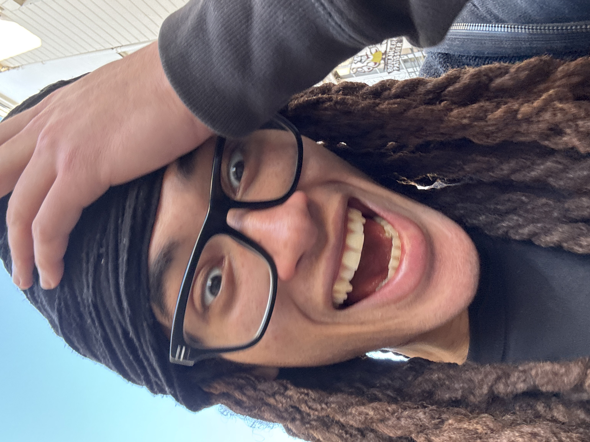 A rotated, close-up selfie captures a person looking directly at the camera with a wide, open-mouthed smile, conveying intense joy or excitement. The person has brown dreadlocks, wears black-framed glasses, and a dark headwrap or beanie. A hand, clad in a dark long-sleeved garment (possibly a hoodie), rests on the side of their head. Their mouth is wide open, showcasing white teeth and a pink tongue.
The background is largely out of focus, but several details are discernible. If the image were rotated upright, a bright blue sky would be visible to the left, suggesting a daytime setting. To the right, a light-colored ceiling with parallel slats or panels and light fixtures can be seen, indicating an indoor or covered outdoor location. Partially visible behind the person's head, on a sign, are the French words "BOULANGER PATISSIER" in a decorative font, a strong clue pointing to a bakery or pastry shop in Paris, France. The overall scene is bright and filled with a sense of playful happiness.