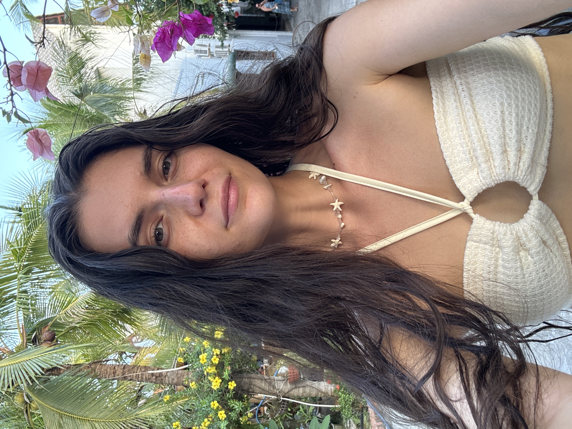 A selfie photograph shows a woman with long, dark brown hair and a light complexion wearing a white crochet bikini top with a thin strap around her neck. She is also wearing a necklace with small, starfish-shaped pendants. Her expression is relaxed and slightly smiling.
The background includes tropical vegetation, such as palm trees and flowering plants with pink and purple blossoms. Yellow flowers are also visible. The scene suggests an outdoor location in a tropical or subtropical climate. The presence of specific tropical plants and architecture suggests that this photograph was taken in Mũi Né, Vietnam.