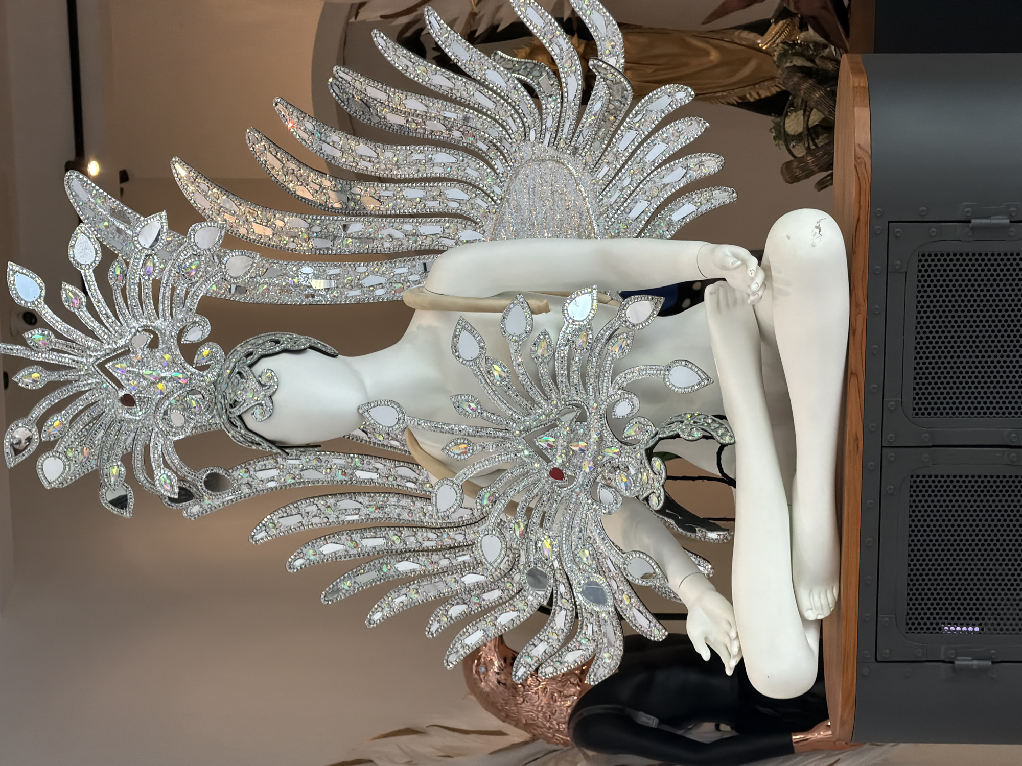 A mannequin wearing an elaborate, silver, feathered headdress and wings adorned with crystals and mirrors is displayed in a seated, meditative pose on top of a dark cabinet with perforated doors. The mannequin is white and smooth, with its legs crossed and hands resting on its knees. Behind the mannequin, to its left, is another mannequin dressed in a black garment with a copper-colored eagle mask. To the right of the main mannequin, other display items and a golden fabric are visible in the background. The scene is indoors, likely a retail or exhibition space, with a neutral-colored wall behind the display. The lighting is even and bright, highlighting the reflective surfaces of the costume. The location is not specified within the image itself; the statement that it is located in Ko Samui District, Thailand, is extraneous information provided by the user.
