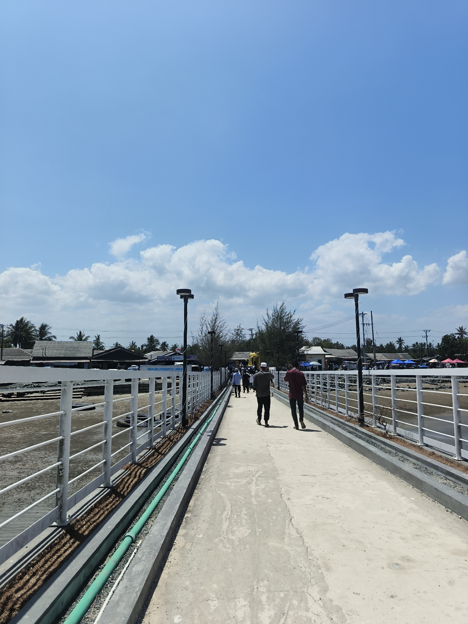 The image depicts a pedestrian walkway, likely in the city of Beringin, Indonesia, on a clear day with scattered white clouds against a bright blue sky. The walkway is paved with concrete and bordered by a white metal railing. Two men are walking away from the camera on the walkway. In the background, there are several buildings, some with dark roofs, and palm trees. There are also some tents visible, suggesting a market or event. A sign that reads "MASJID ZIYADATUL ABRA" is present, indicating a mosque nearby. The walkway appears to be elevated, with an area below covered in sand or shallow water. Two lamp posts with circular fixtures are positioned along the walkway. A green pipe runs alongside the walkway within a concrete channel. The scene suggests a coastal or near-water location.