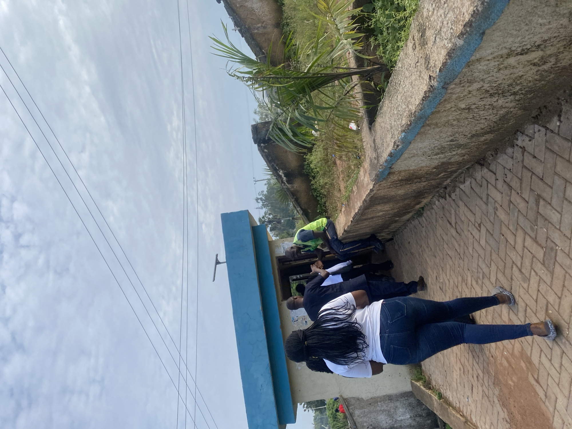 In the city of Rafin Par, Nigeria, a group of individuals gathers near a small, light-colored building with a blue trim. The building appears to be a kiosk or small shelter. A woman with long, dark braided hair, wearing a white t-shirt and dark blue jeans, walks away from the viewer on a paved pathway constructed of rectangular bricks. Behind her, several men are interacting near the entrance of the building. One man in a bright green vest sits on a low concrete wall to the right of the building. The wall has a blue painted edge and is stained with age and weather. Behind the wall, overgrown vegetation and a palm-like plant are visible. Overhead, power lines crisscross the cloudy sky. The overall scene suggests an outdoor public space with a functional, somewhat weathered appearance.