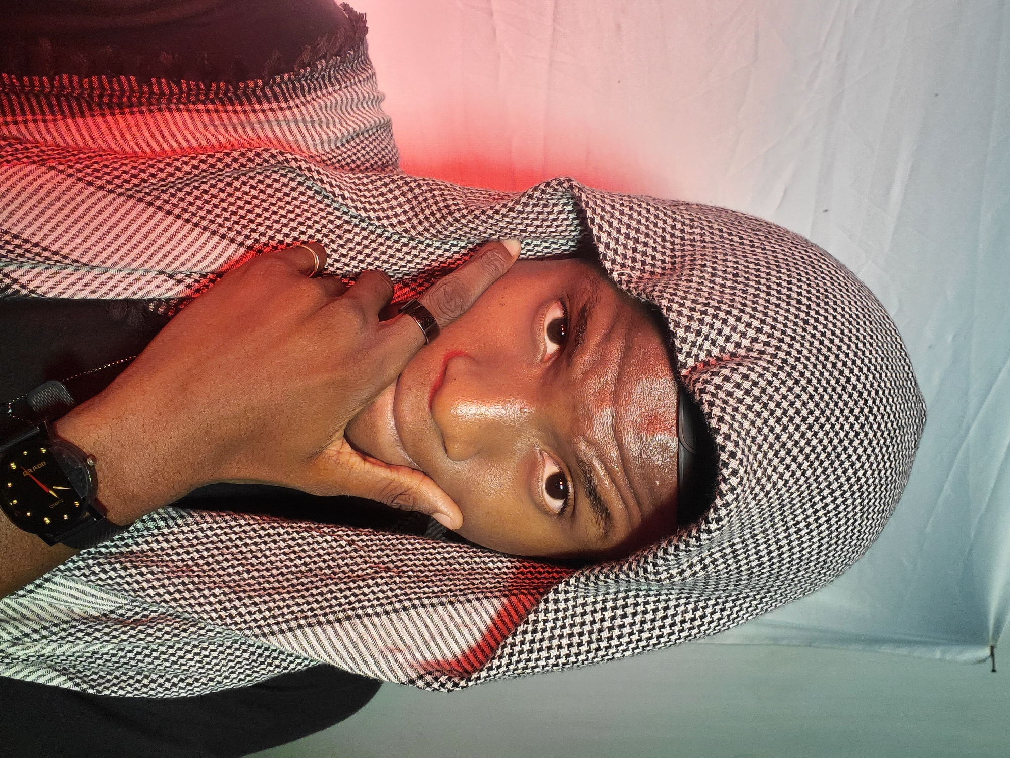 A man with dark skin is angled slightly in a vertical composition. He is wearing a black and white patterned headscarf that covers most of his head. His right hand is raised to his face, with his index finger touching his forehead. He has two rings on his fingers, one silver and one gold. A black wristwatch is visible on his left wrist. The background appears to be a plain white or light gray surface. A red light source illuminates the upper portion of the frame, creating a red hue on parts of the man's face and clothing. The image is taken in an indoor environment. The location is unknown, and there is no information to place the image in Barden Yana, Nigeria.