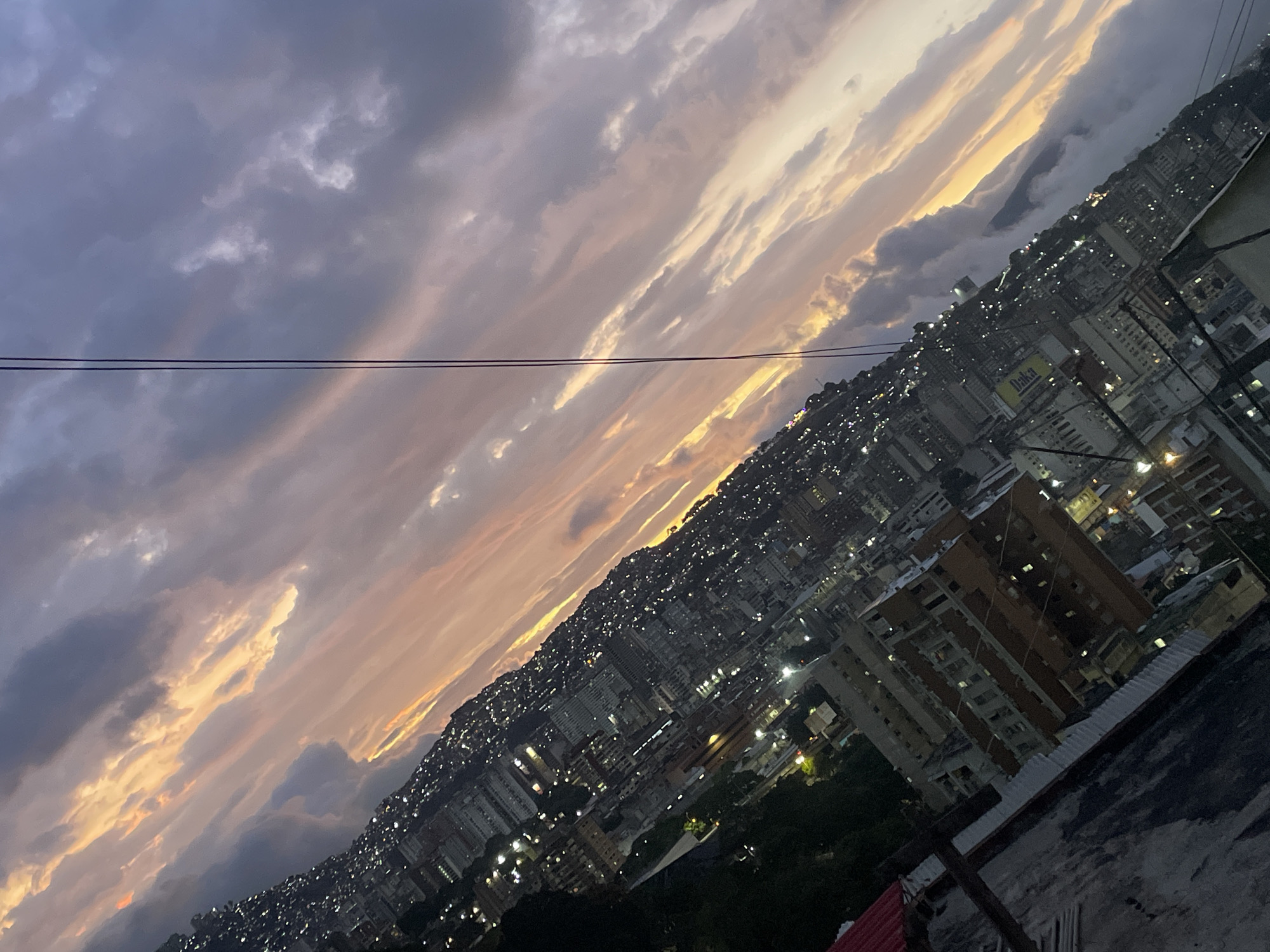 The image depicts a cityscape at dusk, likely taken from a rooftop vantage point. The location is Caracas, Venezuela, recognizable by the densely packed buildings extending up a hillside, characteristic of the city's urban layout. The sky is filled with streaky clouds, transitioning from dark gray to orange and yellow hues near the horizon, indicating a setting or rising sun. Power lines cut across the upper left portion of the sky. The city lights are on, creating a dense, scattered pattern across the buildings. The foreground shows the edge of a rooftop, including various structures and architectural details.