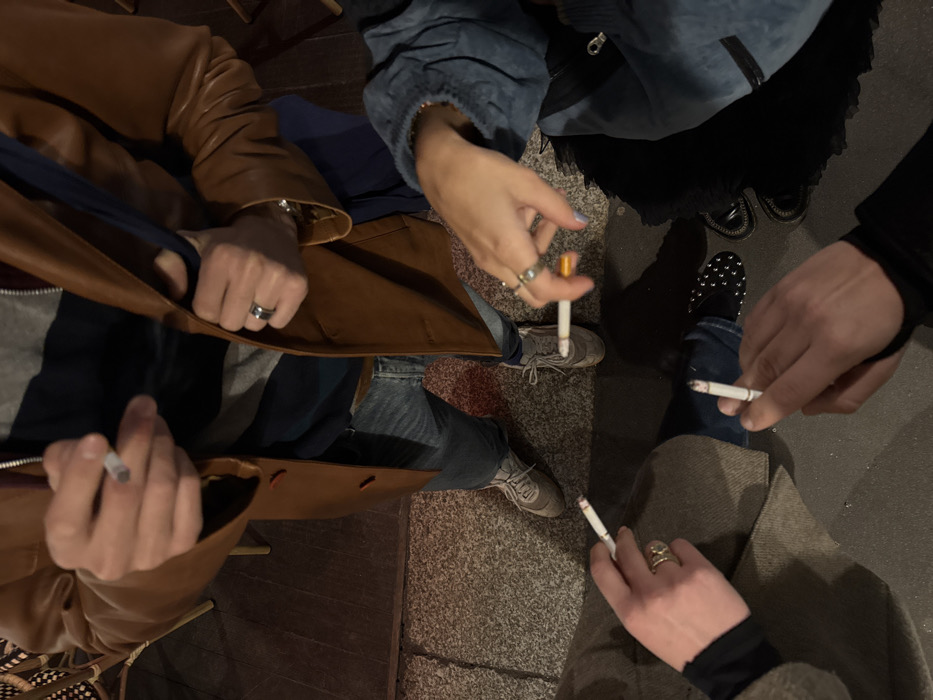 A top-down shot captures four individuals smoking cigarettes, positioned in a circle. The person on the left is wearing a brown leather jacket, while the one next to them sports a denim jacket. The third individual is clad in denim jeans and studded shoes. Lastly, the fourth person is dressed in brown pants and a black turtleneck. The setting appears to be outdoors, with the ground covered in stone tiles. Rings are visible on several hands.