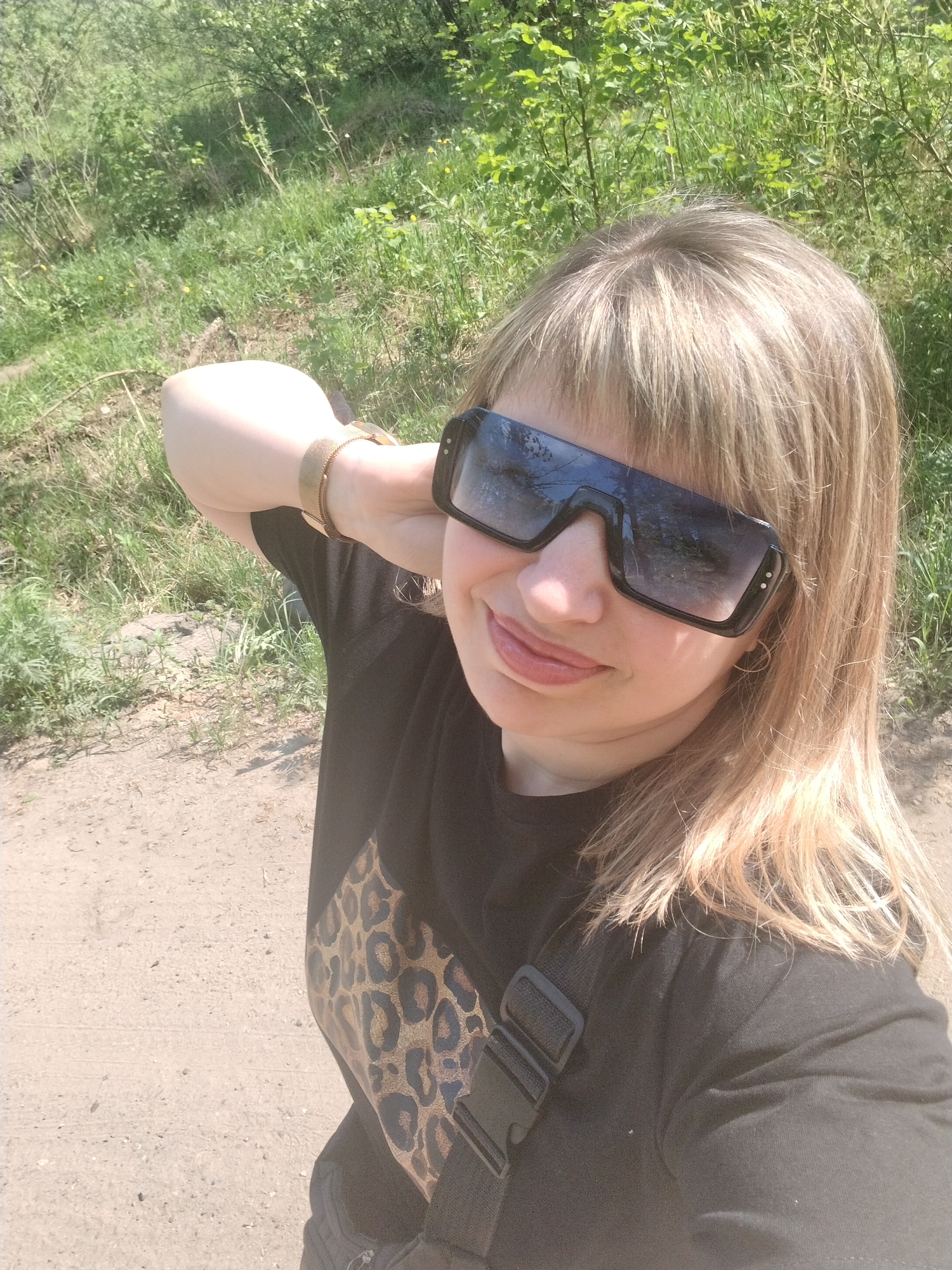 A woman is posing for a selfie outdoors. She is wearing large, dark sunglasses, a black t-shirt with a leopard print design, and a gold-colored bracelet. Her blonde hair is styled with bangs. She is holding her arm up near her head and smiling slightly. In the background, there is green vegetation and a dirt path. The location appears to be a natural area on the outskirts of a city. There is no verifiable information in this image to indicate that this image was taken in Yaroslavl, Russia.