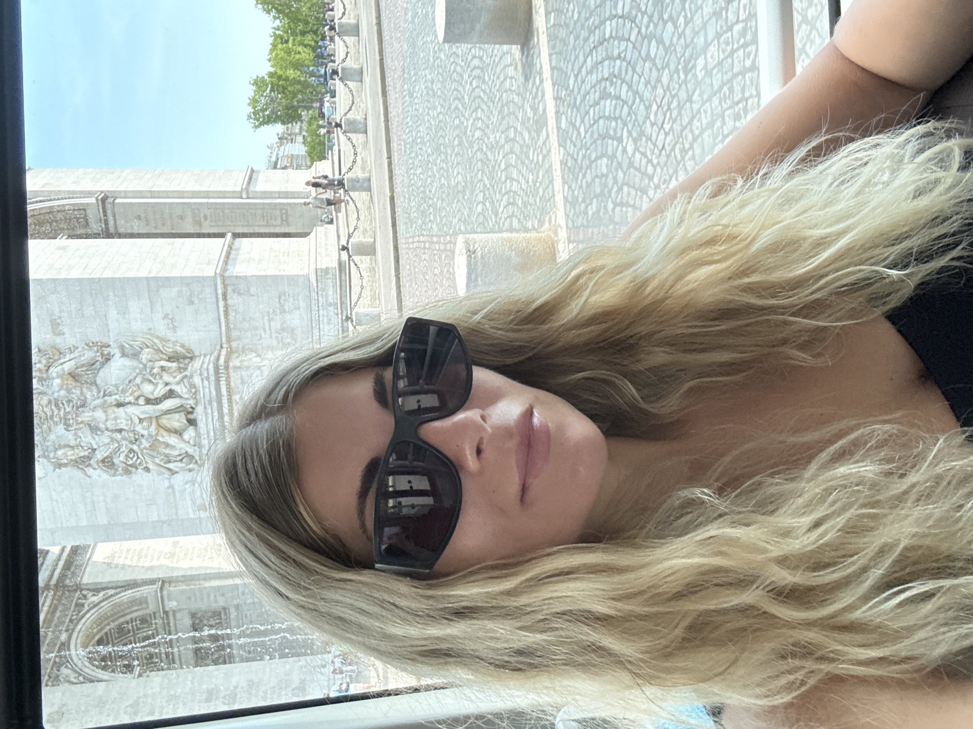 A blonde woman wearing large, dark sunglasses is taking a selfie from a vehicle. She is looking towards the camera with her mouth slightly open. Her long, wavy blonde hair is falling over her shoulders. In the background, the Arc de Triomphe in Paris, France, is visible. The monument, constructed of stone, features ornate sculptures and architectural details. The area around the monument is paved with cobblestones, and there are bollards and chains forming a barrier. Trees and buildings can be seen in the distance, under a bright, clear blue sky. The scene appears to be a sunny day.