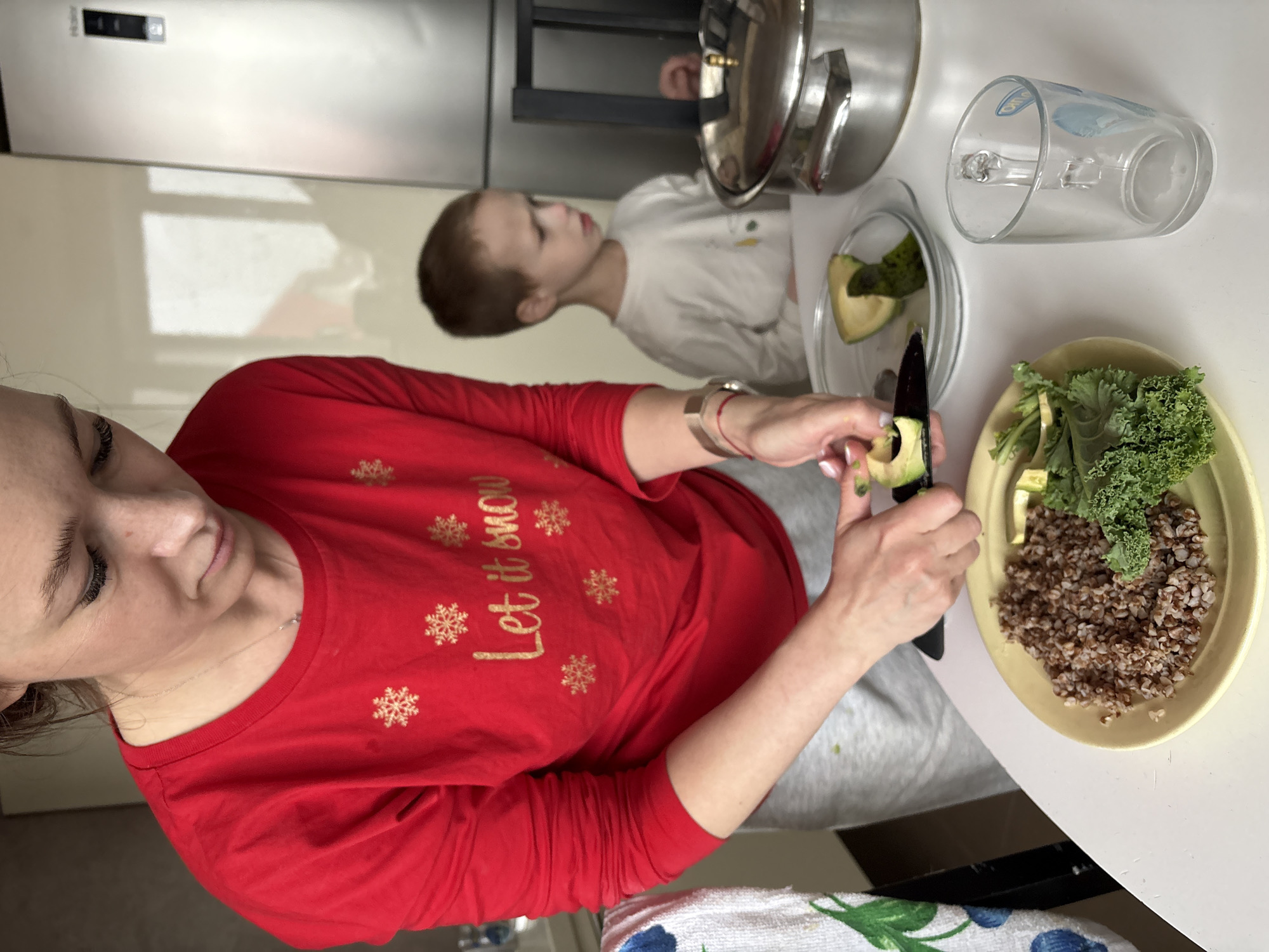 A woman and a young child are captured in a domestic scene, likely in a kitchen or dining area in Ulyanovsk, Russia. The image is taken from an overhead perspective, focusing on food preparation at a white table.
The woman, positioned in the lower-left portion of the frame, is wearing a festive red long-sleeve shirt decorated with gold snowflakes and the phrase "Let it snow." She is actively preparing food, holding an avocado in her left hand while using a black-handled knife in her right to peel or cut it. Her hands feature a light-colored manicure. She appears focused on her task.
On the white table in front of her, there's a yellow plate containing a serving of cooked buckwheat and fresh green kale. Another clear plate holds cut avocado pieces. A transparent glass and a silver pot with its lid are also visible on the table.
To the right and slightly in the background, a young child with short brown hair is observing the woman's activity. The child is wearing a white long-sleeve shirt and is looking towards the food being prepared.
In the background, a silver Haier refrigerator is partially visible against a light-colored wall. Reflections on the refrigerator suggest a bright, daytime setting. A towel with a white and blue floral pattern is seen in the immediate foreground, resting on what appears to be a chair or lower surface. The overall scene depicts a calm, everyday moment of meal preparation.