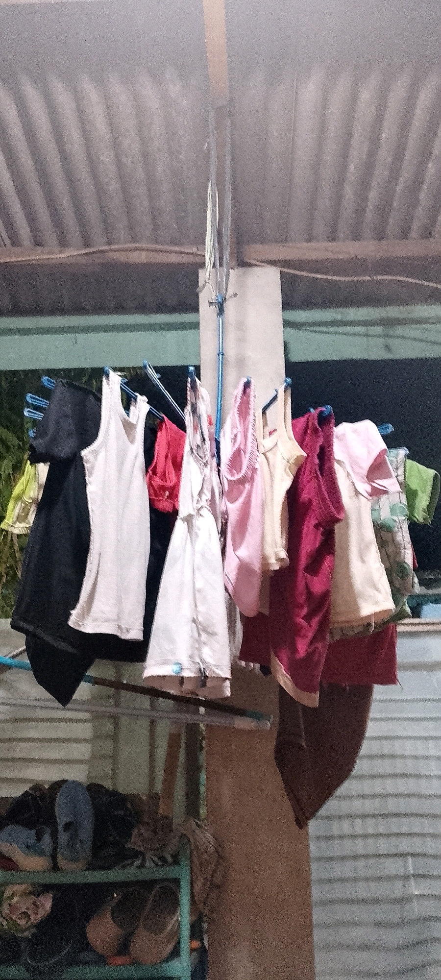 This image depicts a clothesline outdoors, likely in the city of Satriamekar, Indonesia, under a corrugated metal roof. A variety of clothing items, including tank tops and t-shirts in pastel colors and a darker hue, are hung on blue hangers. The clothing appears to be damp, suggesting it is drying. Beneath the clothesline, a green shelf unit holds several pairs of shoes, including sandals and closed-toe shoes. The background is dark, hinting at it being either dusk or night. The overall scene suggests a domestic setting where laundry is being air-dried.
