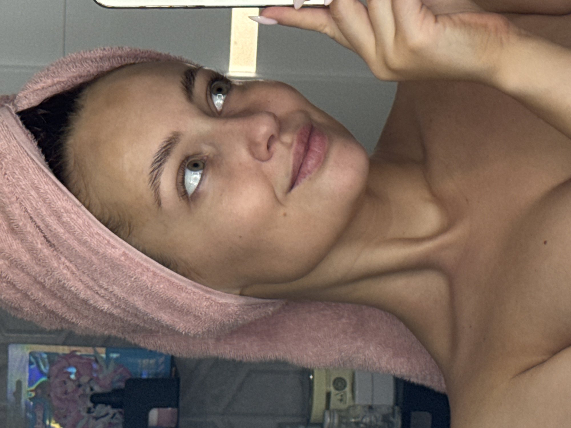 A woman with fair skin and light brown hair wrapped in a pink towel is holding a smartphone above her head, presumably taking a selfie. She is facing the camera with a slight upward gaze. The background is blurry, but parts of a cluttered room with items like a box, a bottle, and a power outlet are visible. The location in Chaikovsky, Russia, cannot be determined from the image alone.