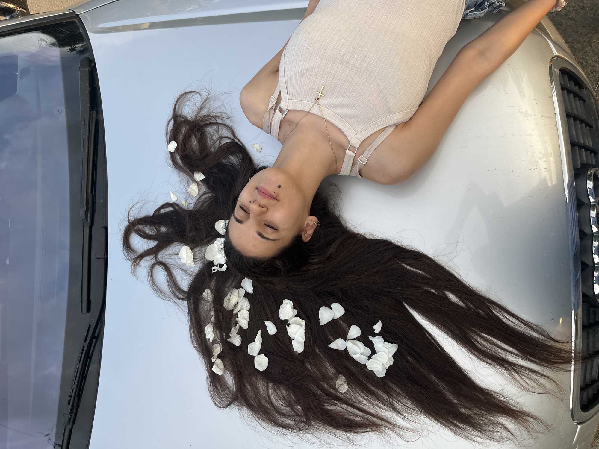 A young woman with long, dark brown hair lies serenely on her back across the silver hood of a car. Her eyes are closed, and a faint smile plays on her lips, suggesting a peaceful or relaxed state. White flower petals are scattered throughout her hair and on the car's surface around her head, creating an ethereal and gentle aesthetic.
She is dressed in a light beige or cream-colored ribbed tank top or bodysuit, featuring thin straps. A delicate gold chain with a cross pendant hangs around her neck, resting on her chest. Her long hair is spread out behind her head and extends down the hood of the car, with some strands flowing towards the front grille.
The car appears to be a silver or light grey model, possibly a sedan or SUV, with part of its windshield, wipers, and front grille visible. The photo is taken from above, looking down on the woman and the car. The setting is outdoors, under clear daylight, indicated by the bright illumination and soft shadows. The overall scene evokes a calm and artistic mood. No discernible text is visible in the image. The location context provided is Lozarevo, Bulgaria.