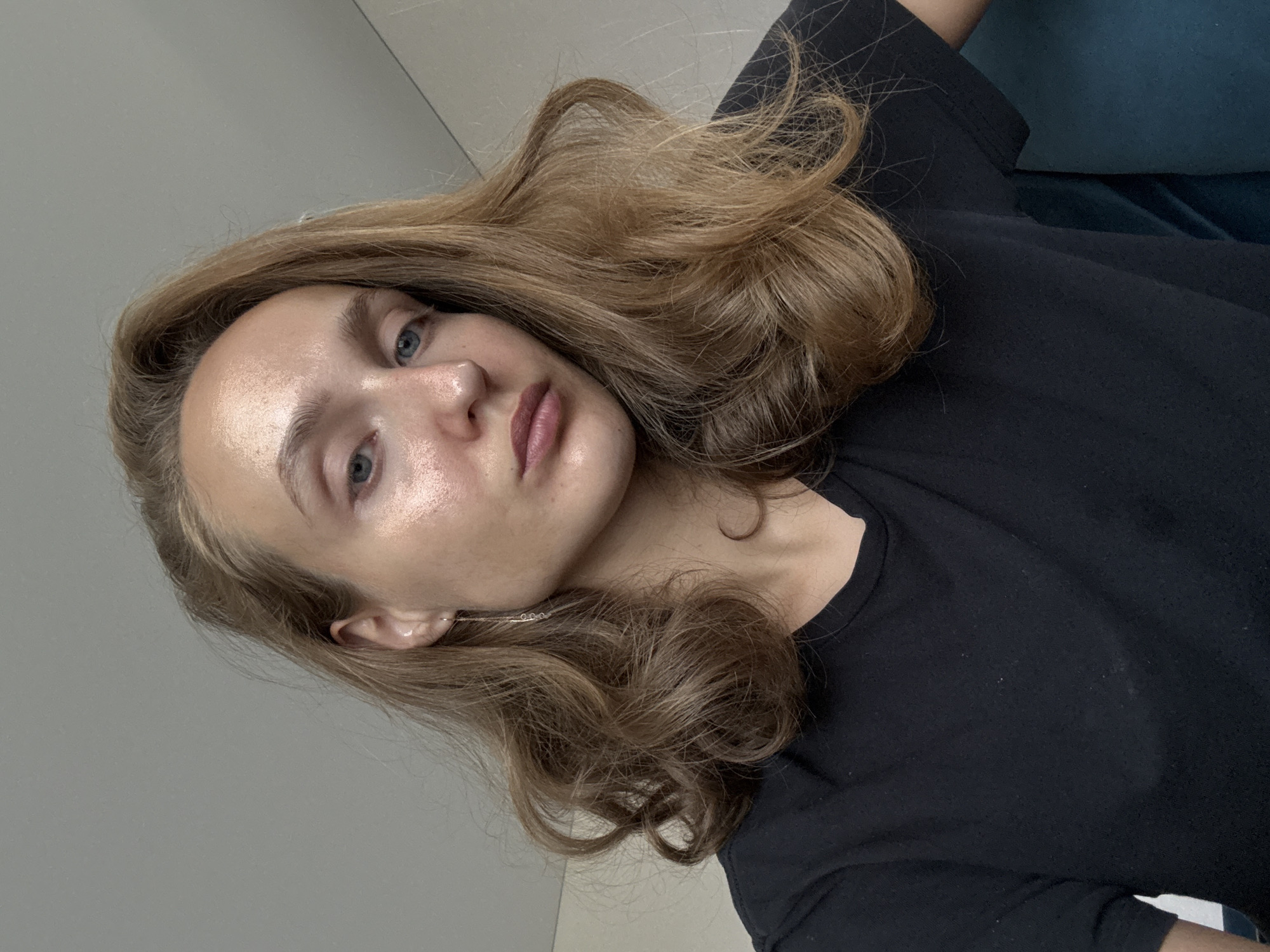 A young woman with shoulder-length, wavy, light brown hair is shown in a close-up, slightly angled selfie. She is wearing a plain black t-shirt. Her face is illuminated, highlighting her blue eyes, fair skin with a subtle glow, and light makeup on her lips. She has a single, delicate dangle earring in her left ear. The background is a plain, light grey wall. There is no indication of the location being in the city of Подшивалово, Russia.