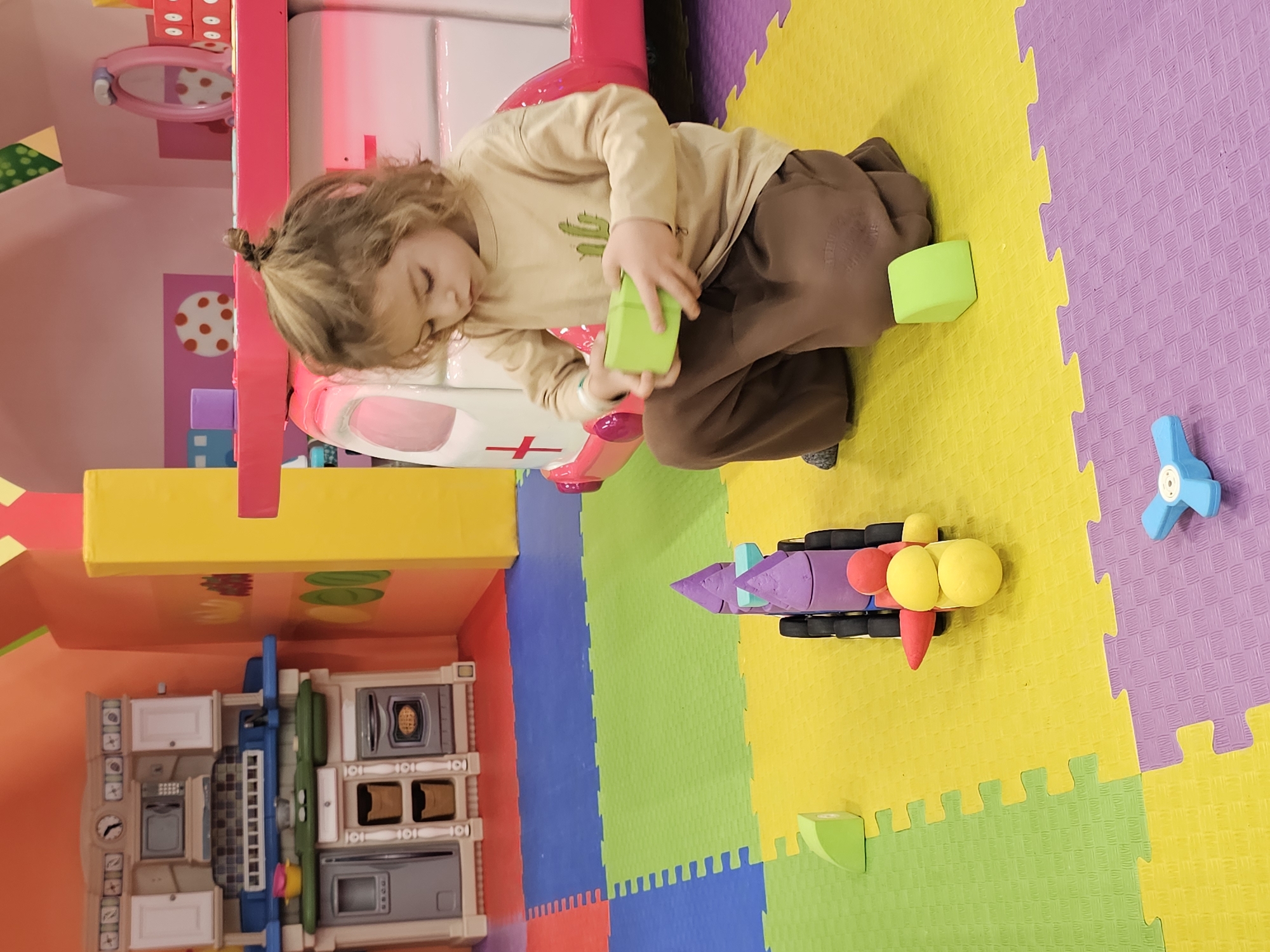 A child with curly blonde hair, styled in a small ponytail, is kneeling on a playmat composed of interlocking foam tiles in various colors (purple, yellow, green, red, and blue). The child is wearing a light beige long-sleeved shirt with a cactus graphic and brown pants. They are holding two green foam blocks.
To the child's left is part of a pink and white play structure, which includes a play ambulance. To the lower-left, there's a toy kitchen set with appliances.
To the right of the child, there is a toy rocket and a blue propeller-shaped toy on the playmat.
The location is identified as Khimki, Russia.