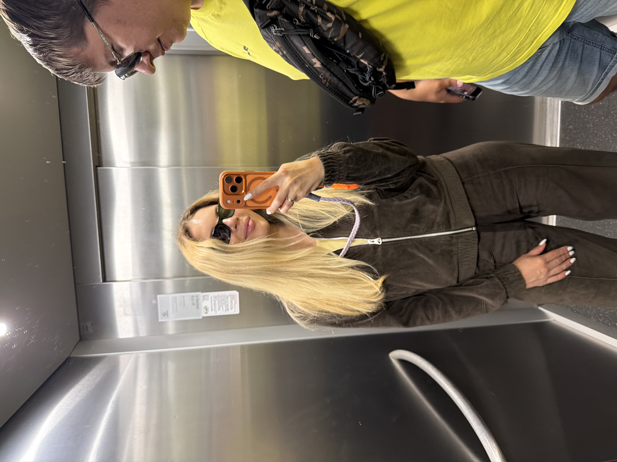 A mirror selfie is taken in an elevator, showing two people. A person in a yellow shirt, denim shorts, and a camouflage-patterned bag is reflected at the top of the mirror. Below, a person with long blonde hair, sunglasses, and a brown velvet tracksuit takes the selfie with an orange phone. The elevator's interior consists of a stainless steel wall with a white safety information sticker. The presence of a person and the style of clothing suggest a recreational setting. The location, Budva, Montenegro, is not visually determinable from the image.