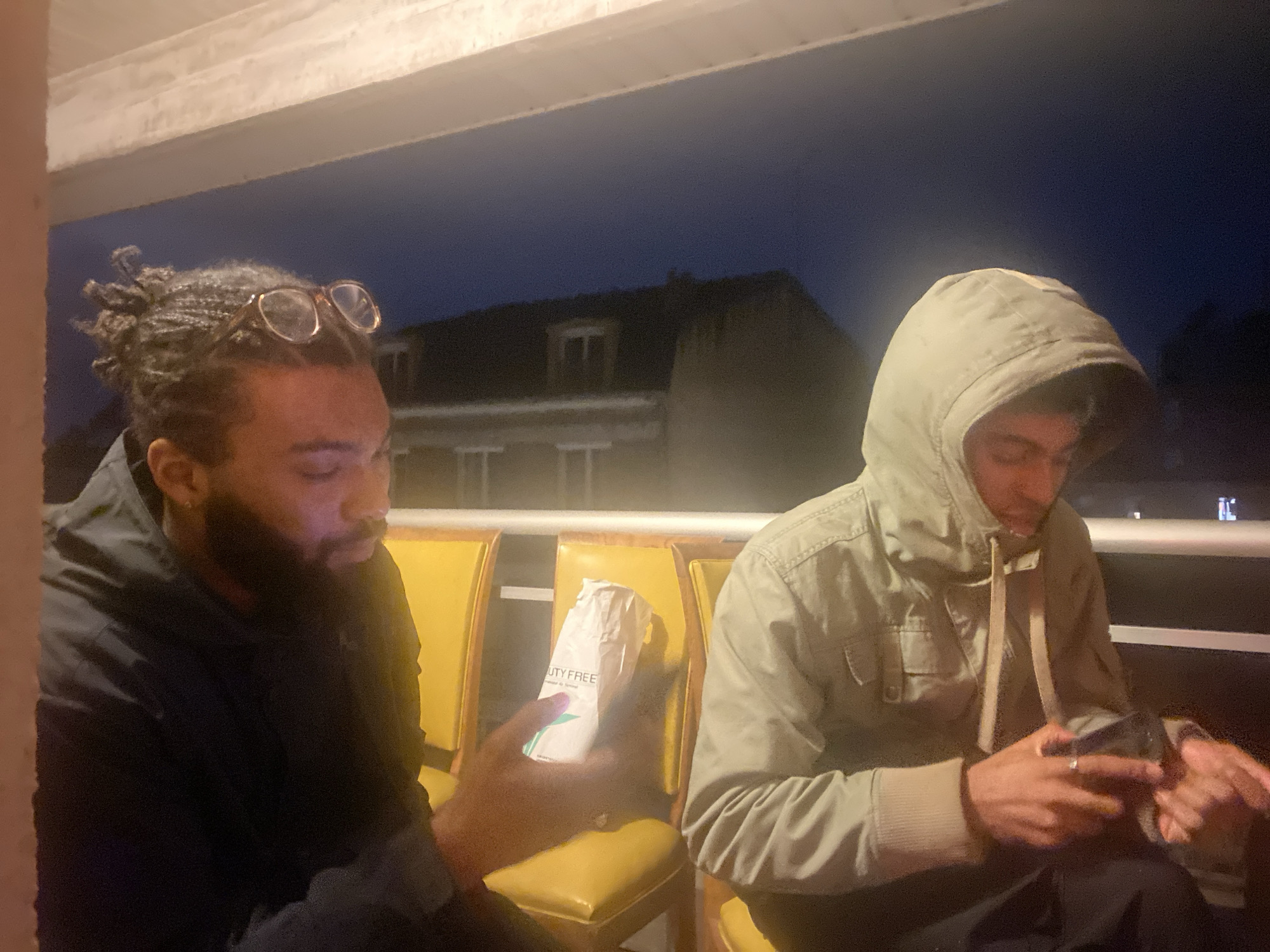 Two men are seated in yellow chairs under an overcast sky, likely in Montreuil, France. The man on the left has dark hair styled in dreadlocks with glasses resting atop his head. He has a dark beard and is wearing a black hoodie. He's holding a white paper bag with the words "DUTY FREE" printed on it. The man on the right is wearing a hooded light-colored jacket. He has his hood up and is looking down at a cell phone in his hands. They are both sitting outside near a building.