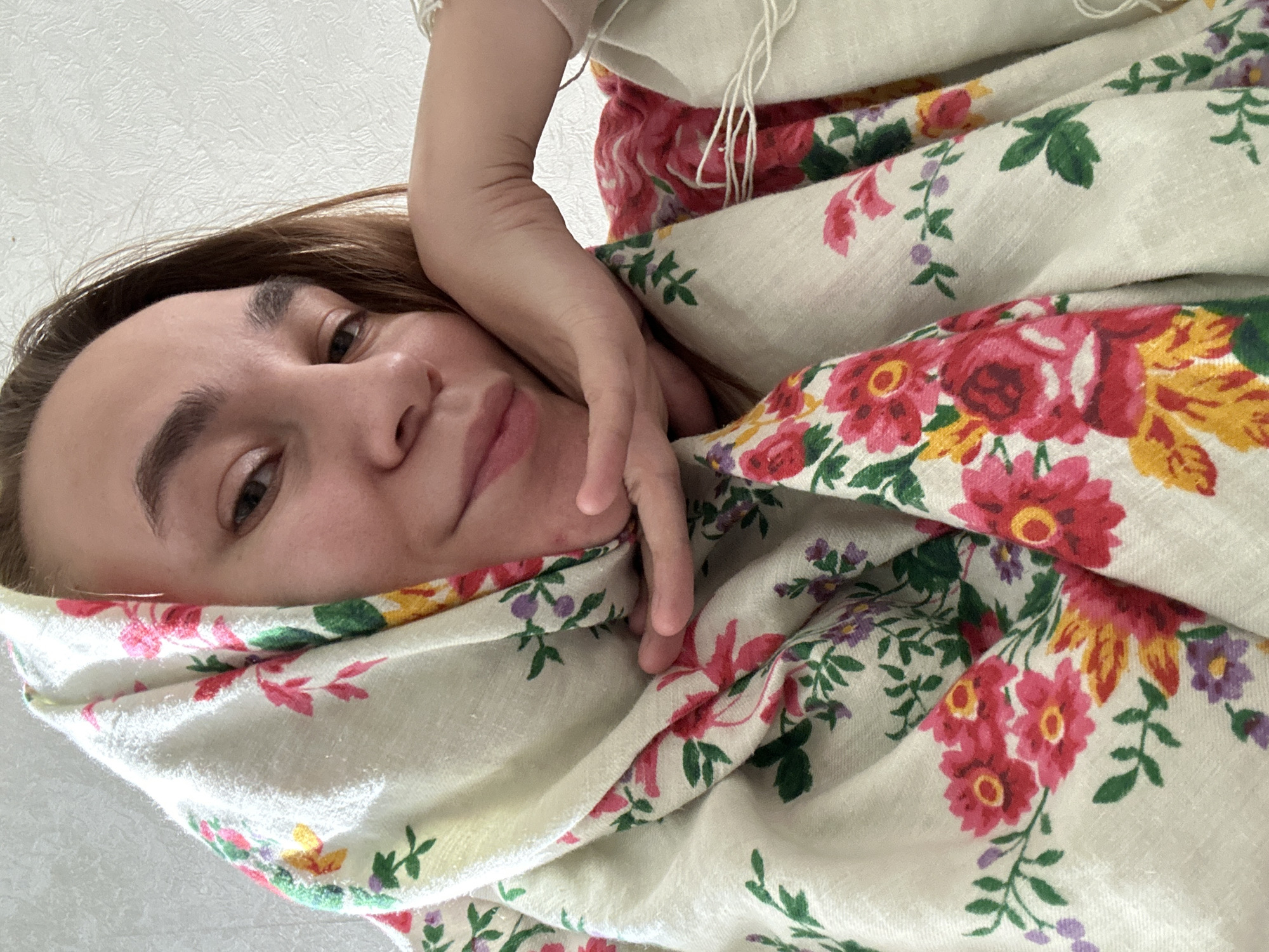 The image presents an overhead, close-up view of a woman, likely taking a selfie or being photographed from above. Her face, characterized by light brown hair, dark eyes, and a soft, neutral expression bordering on a faint smile, is positioned in the lower left quadrant, looking directly at the viewer.
She is partially enveloped and surrounded by a voluminous, light-colored fabric adorned with a bold floral pattern. This pattern features clusters of large red, pink, and yellow flowers, interspersed with green leaves and smaller purple elements, all set against a cream or off-white background. The fabric creates gentle folds around her, and her right arm and hand are visible, resting near her face and tucked into the material. Another segment of light-colored fabric, possibly a garment or another scarf, with visible tassels, can be seen in the upper right of the frame.
The background consists of a plain, textured white surface, suggesting an indoor setting, likely a wall or ceiling. The lighting appears bright and even, indicating daytime. The overall impression is one of comfort and relaxation. No specific location cues are discernible within the image, but the provided context for the setting is Uva, Russia. There is no visible text in the media file.
