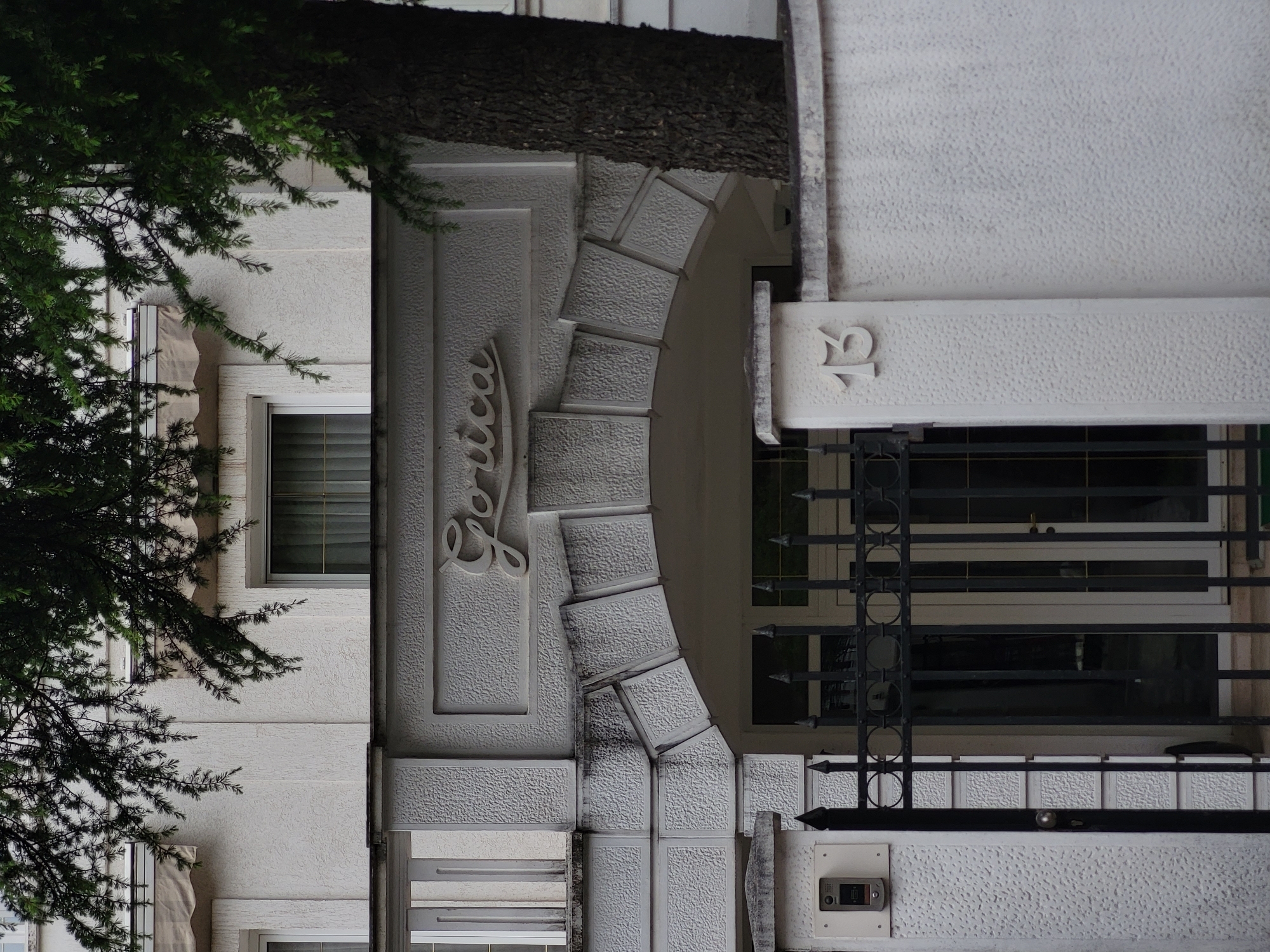 The image displays the exterior of a white, multi-story building, likely an apartment complex or hotel, characterized by its textured stucco facade. A prominent arched entrance serves as a central feature, adorned with decorative, textured block-like voussoirs.
Above the archway, a raised, stylized sign spells out "Gorica" in a flowing script. To the right of the arch, the number "13" is embossed on a flat section of the wall. The entrance is secured by a black wrought-iron gate or railing, featuring ornate circular patterns and pointed finials. Below the gate, an intercom system is mounted on the wall.
Several windows with internal blinds or shutters are visible on both sides of the archway. The left side of the building is partially obscured by the lush green branches and needles of a tree or large bush. A dark tree trunk is also visible overhead, casting some shade on the building. The lighting suggests it is daytime, but appears soft and diffused, indicating an overcast sky or a shaded area. No activity is taking place. The architectural elements, such as the white stucco and wrought iron, are consistent with structures found in Mediterranean and Balkan regions, aligning with the location context of Budva, Montenegro.